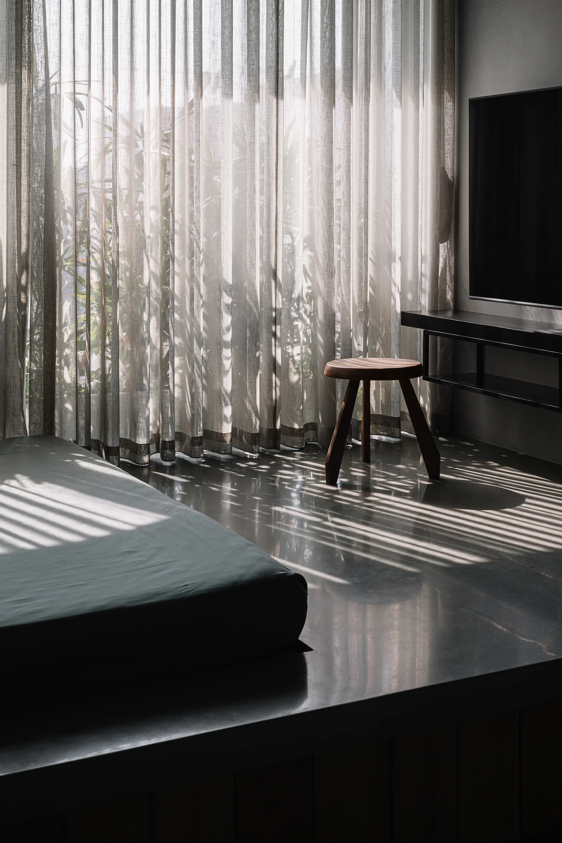 A low platform bed and a wooden accent stool stand on polished concrete floors in a minimalist bedroom with sheer light-filtering curtains.