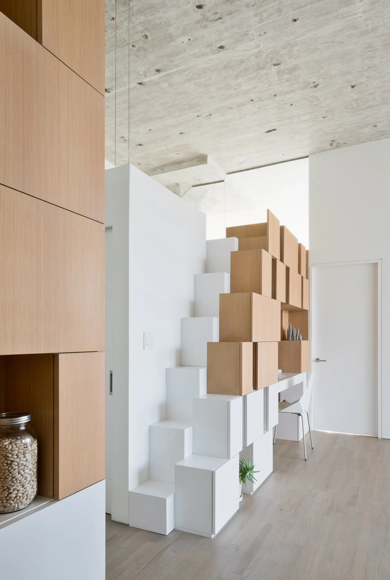 A minimalist white modular staircase with integrated wood-toned storage cubes next to a small built-in desk under a raw concrete ceiling.