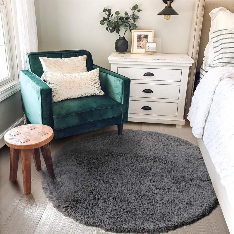 A cozy bedroom corner featuring an emerald green velvet armchair, a distressed white nightstand, and a plush round grey shag rug.