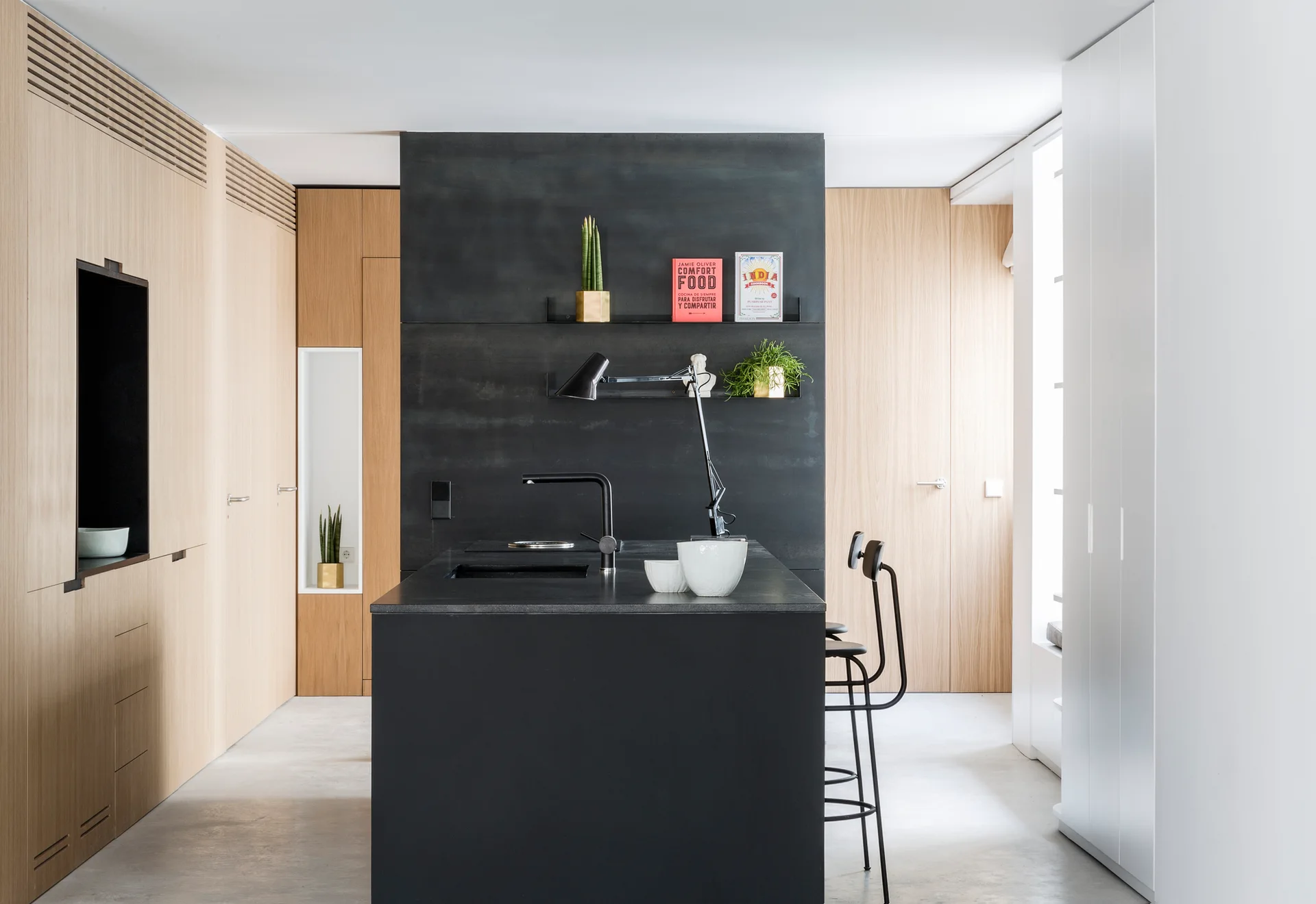 A modern minimalist kitchen with a central matte black island, built-in light wood cabinetry, concrete flooring, and a black accent wall.