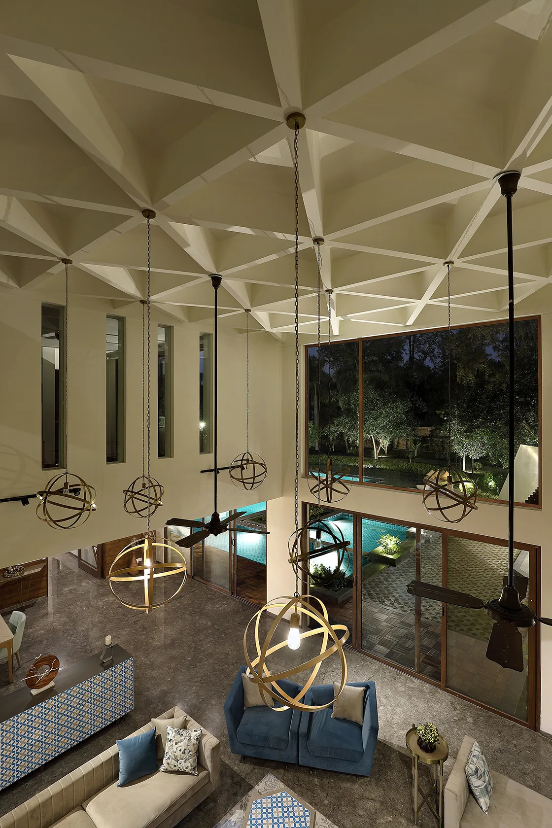 A high-angle view of a double-height living room featuring a geometric grid ceiling, spherical pendant lights, and large windows opening to a pool.