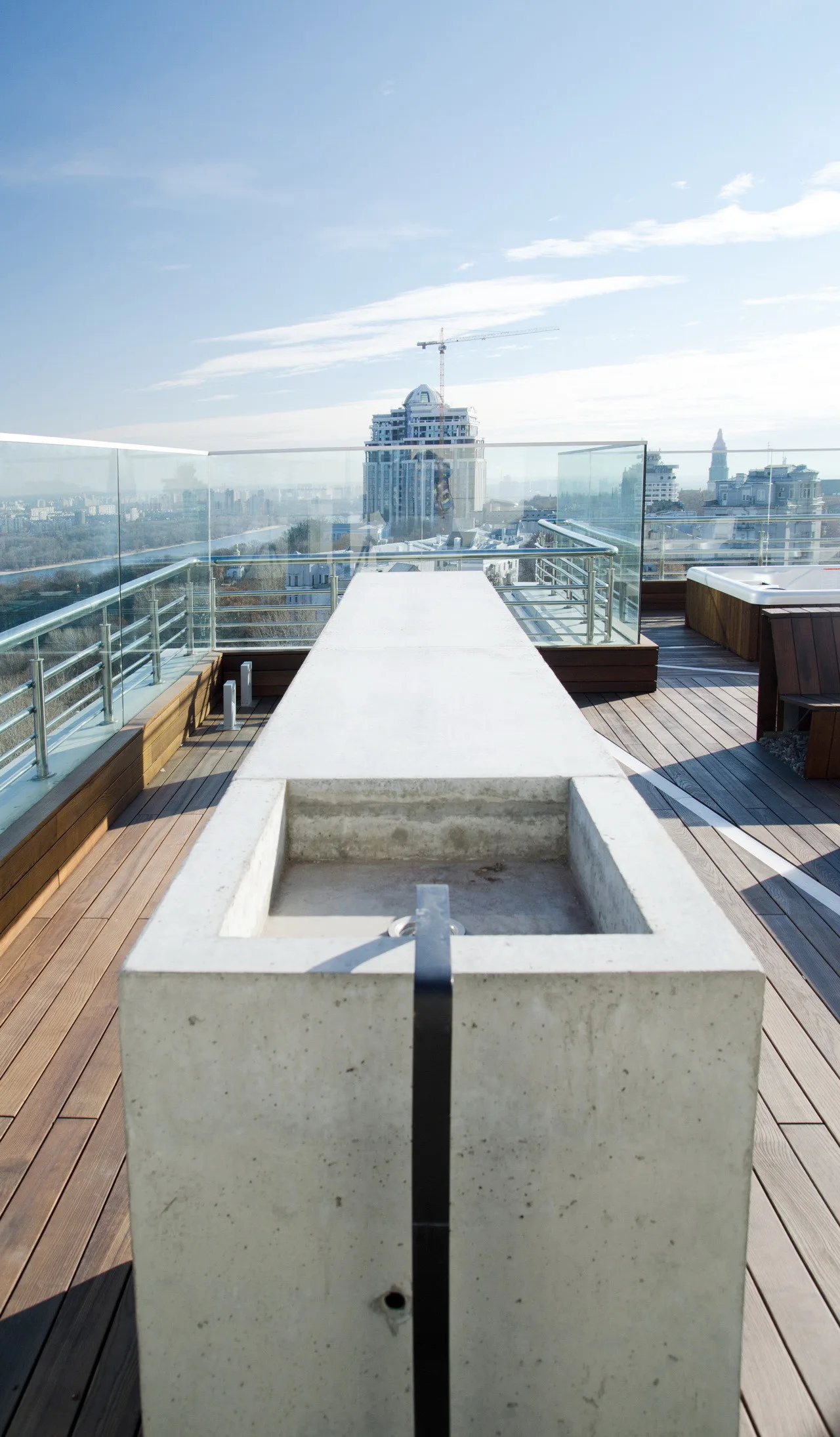 A long grey concrete counter stands on a wooden deck on a rooftop terrace enclosed by glass railings overlooking a city.