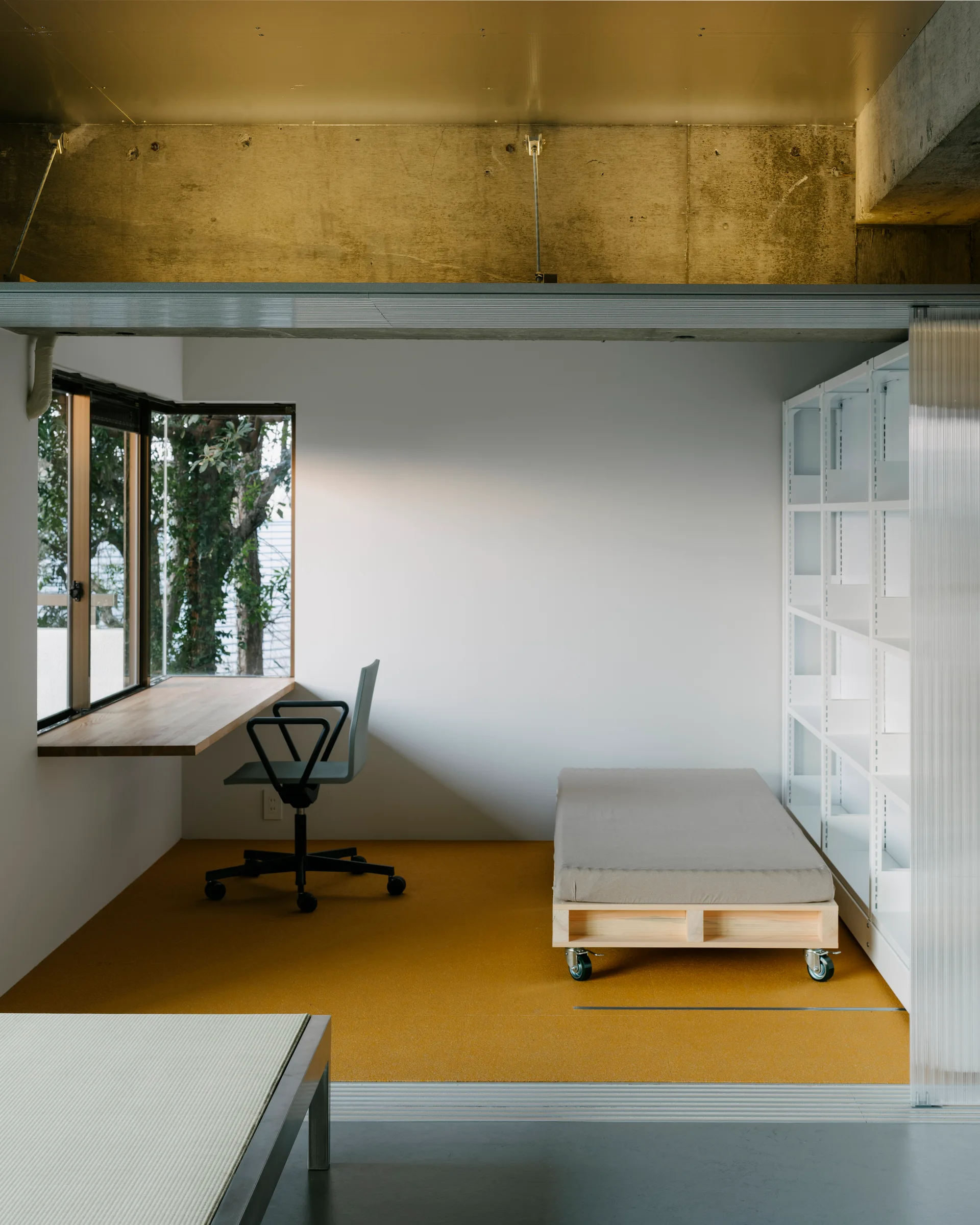 A minimalist study room featuring a mustard yellow floor, a wall-mounted wooden desk, and a low platform bed on wheels.