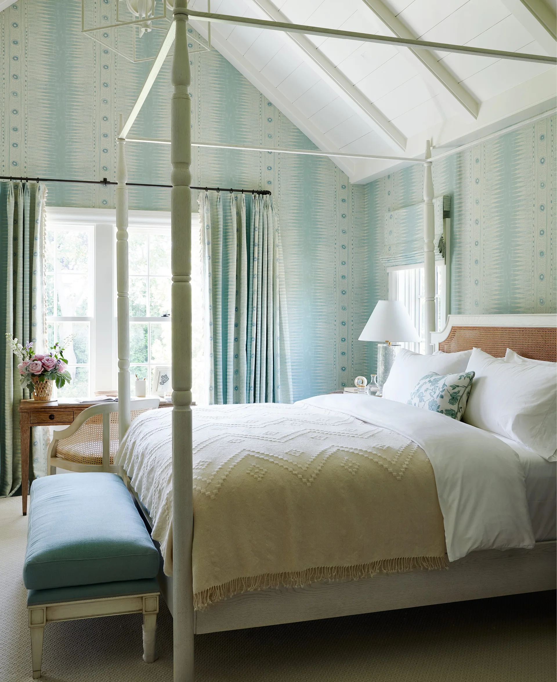 A light blue bedroom features a tall cream canopy bed, matching patterned wallpaper and curtains, a vaulted ceiling, and an upholstered bench.