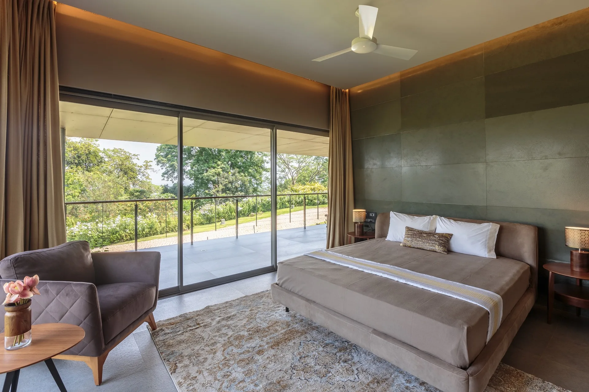 A modern bedroom featuring a brown upholstered bed against an olive green paneled wall next to large sliding glass doors.