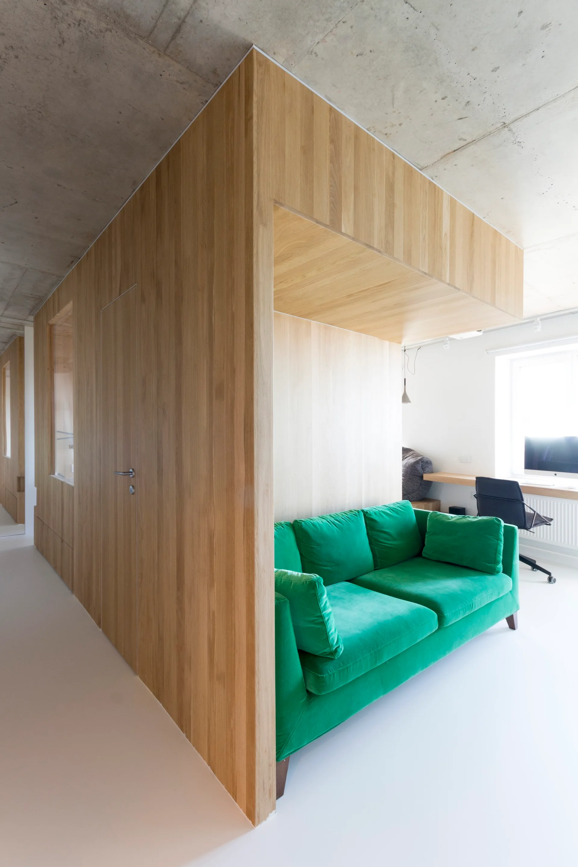 A green velvet sofa is tucked into a light wood paneled wall niche beneath a raw concrete ceiling.