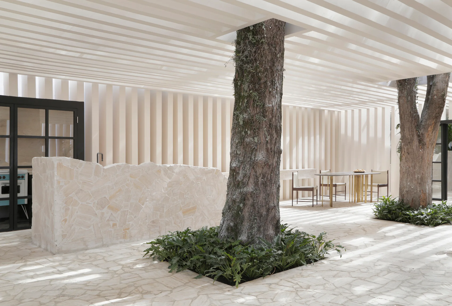 An outdoor cream-colored patio features mature trees growing through a stone tile floor beneath a white slatted ceiling.