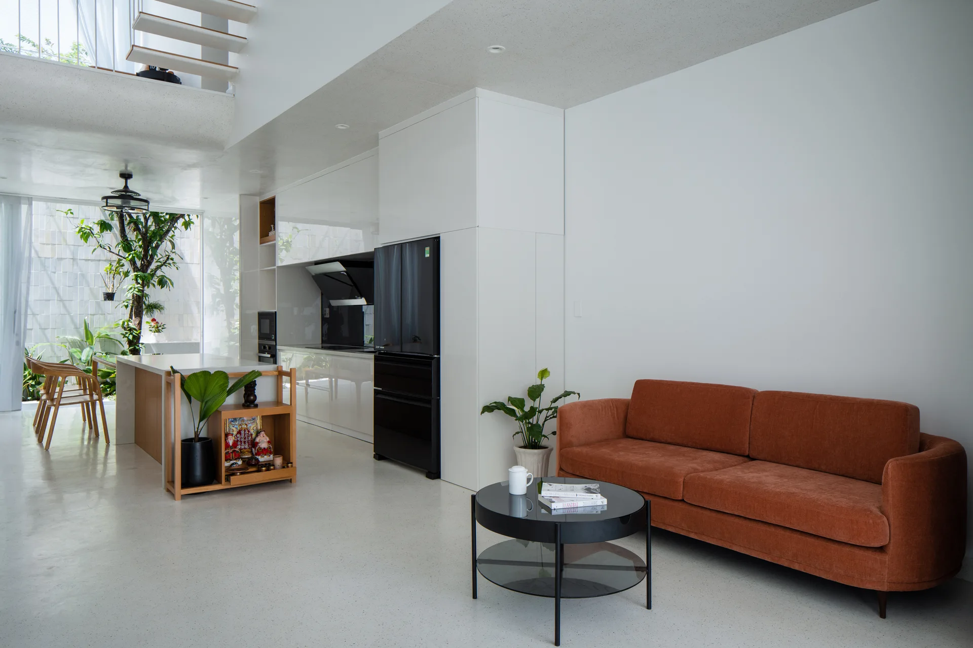 A bright open-plan living room and kitchen featuring a rust orange sofa, a black glass coffee table, and sleek white cabinetry.