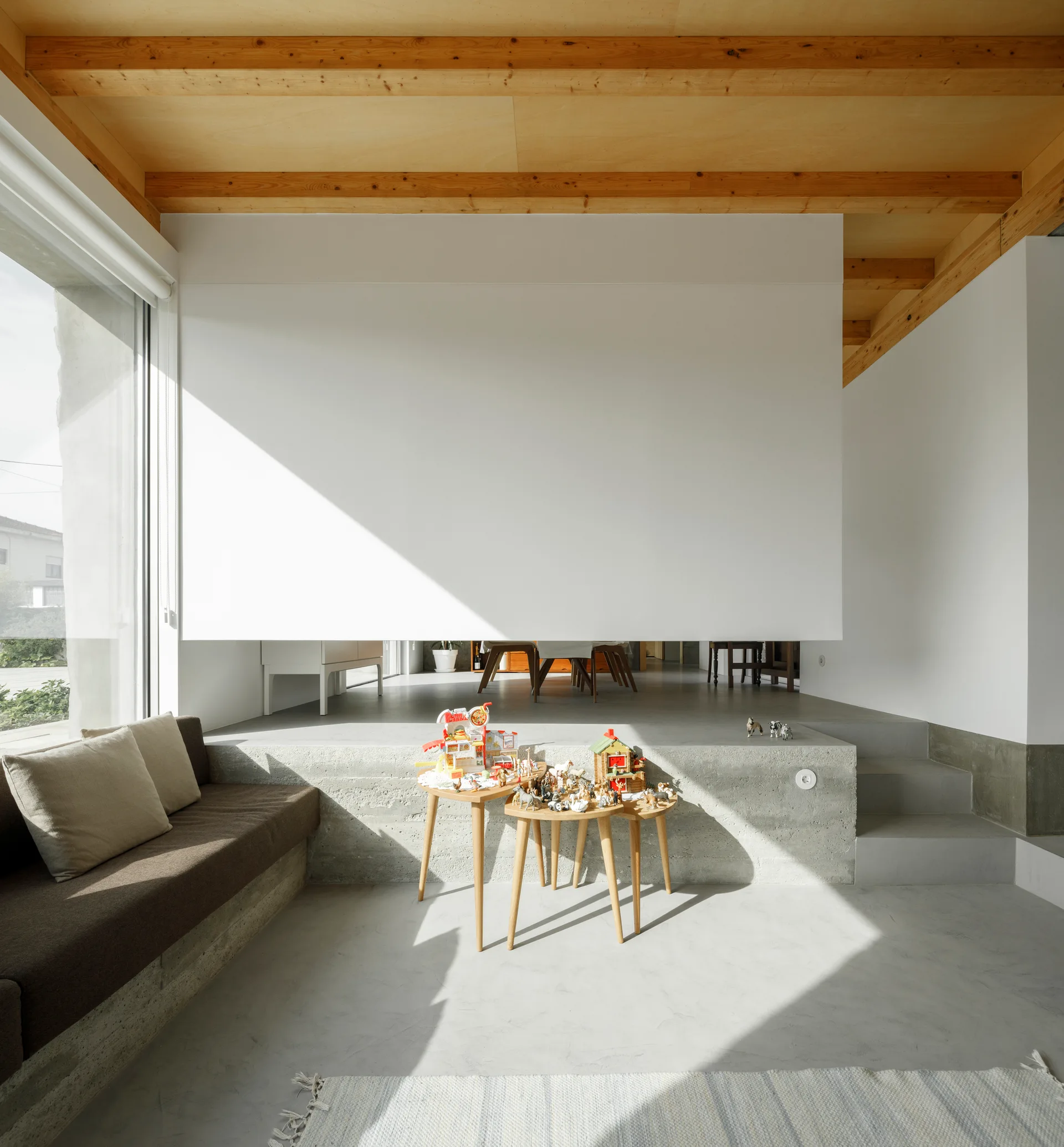 A modern split-level living room features concrete steps, an upholstered concrete bench, and a warm exposed timber ceiling with wooden beams.