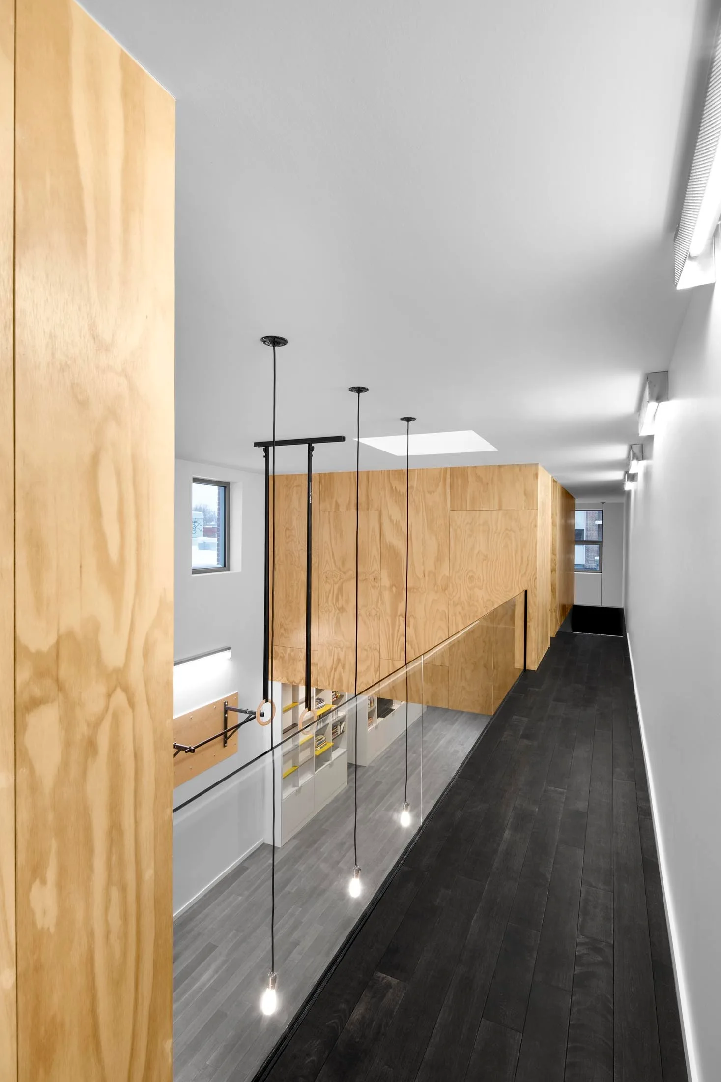A modern mezzanine corridor featuring light plywood wall paneling, dark wood flooring, and a transparent glass railing with hanging pendant lights.