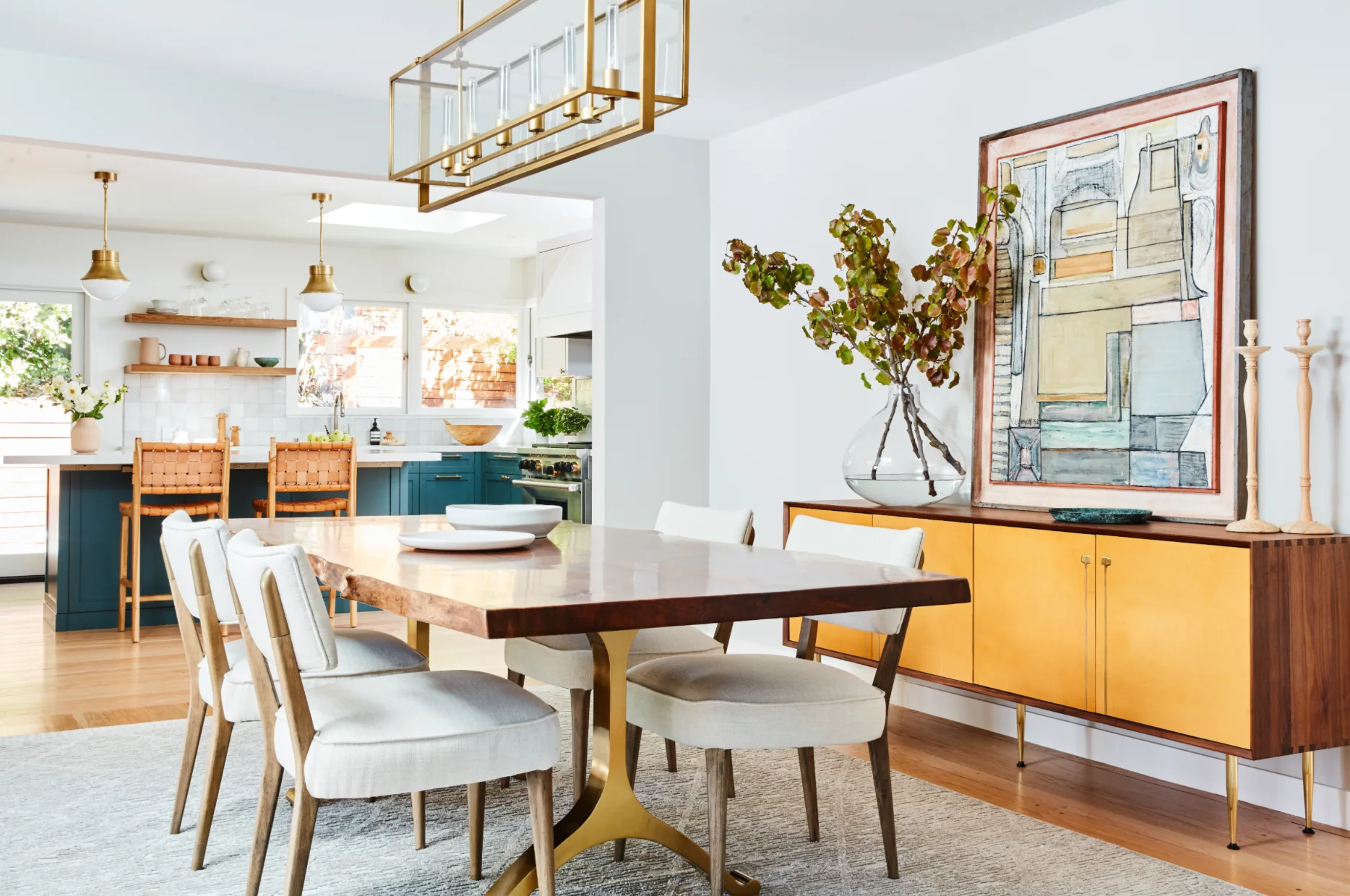 A bright dining room features a live-edge wooden table, white upholstered chairs, a yellow sideboard with abstract art, and a view into a teal kitchen.