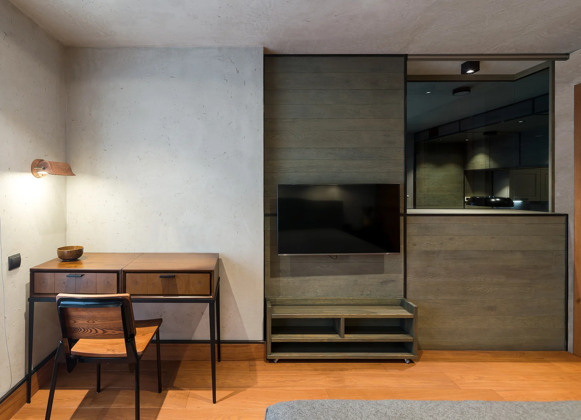 A modern bedroom workspace featuring a wooden writing desk, a matching chair, a wall-mounted TV on a wood-paneled accent wall, and textured concrete.