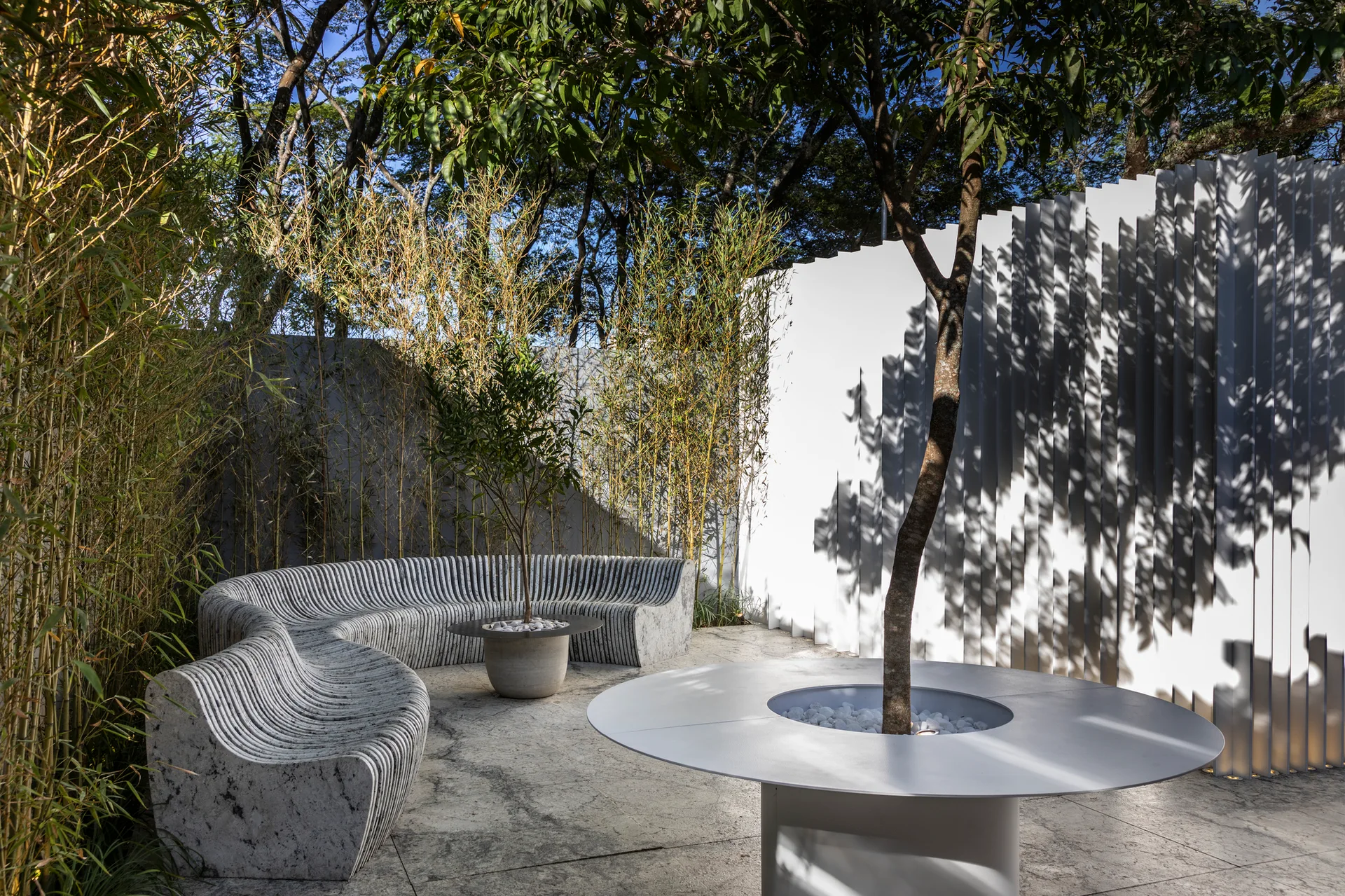A stone patio features a wavy grey stone bench, a circular white table built around a tree, and a white vertical slat wall backdrop.