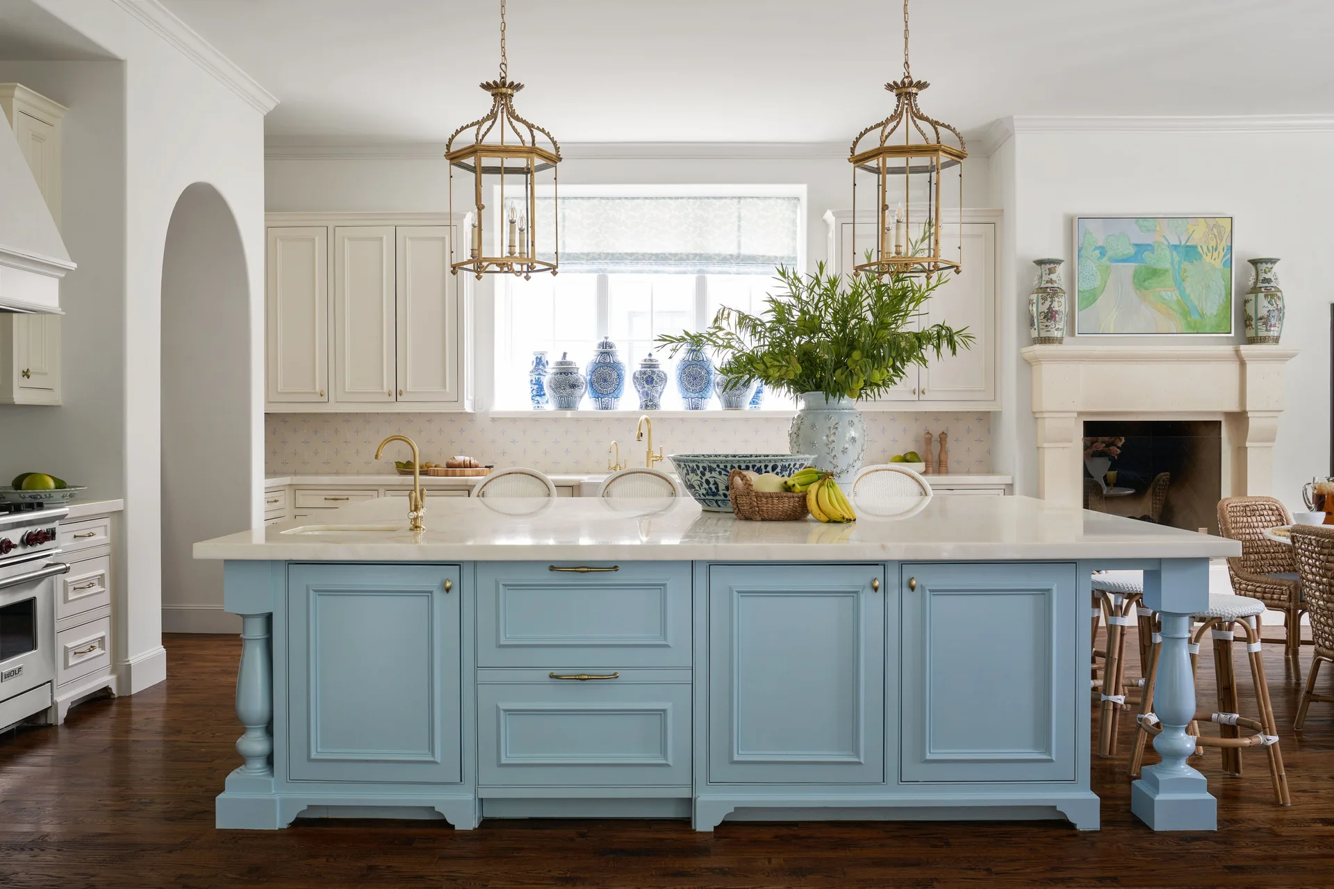 A white kitchen features a light blue island with a marble countertop, brass lantern pendants, and woven bar stools.