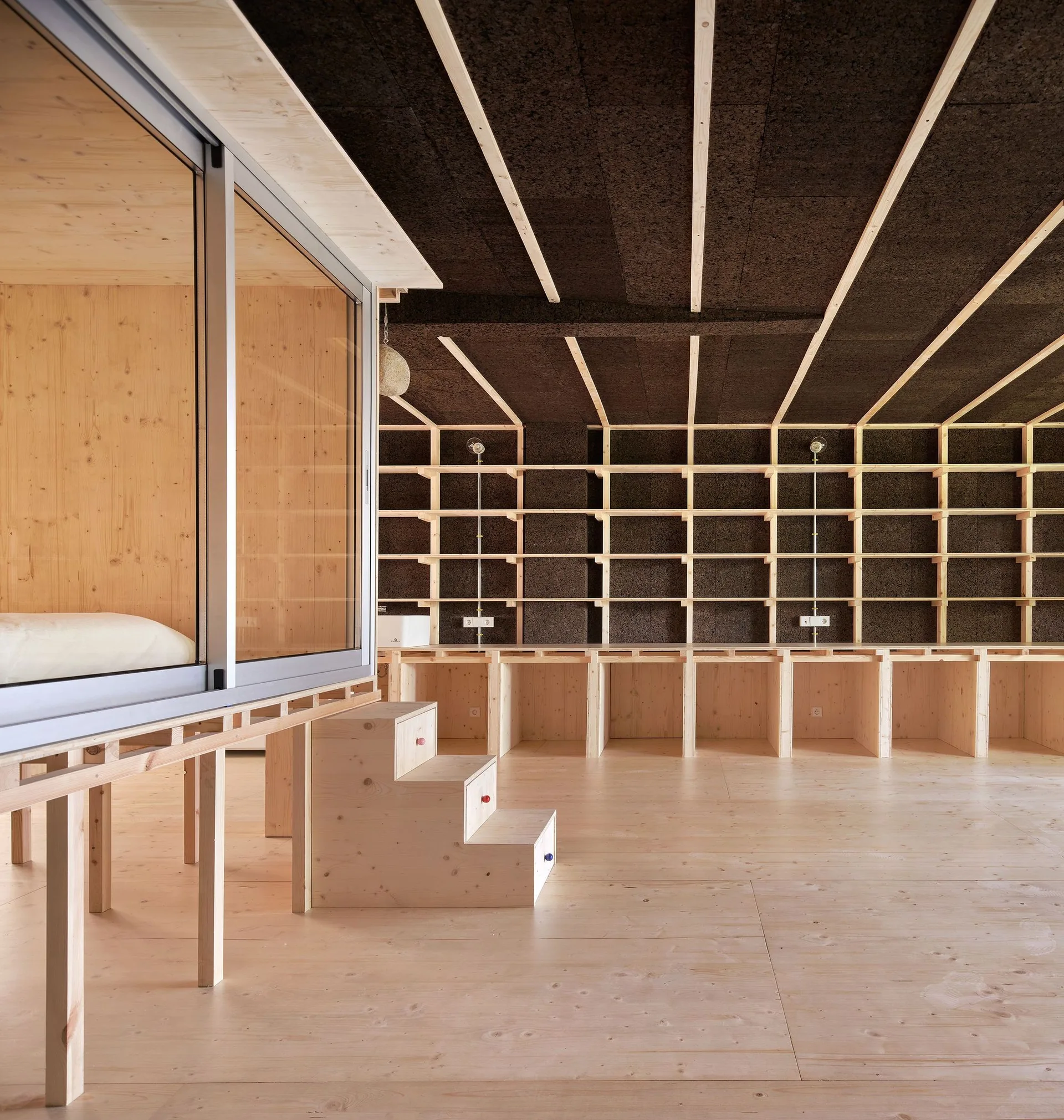 A minimalist room featuring an elevated sleeping platform with sliding doors next to a wall of light wood shelving and a dark cork ceiling.