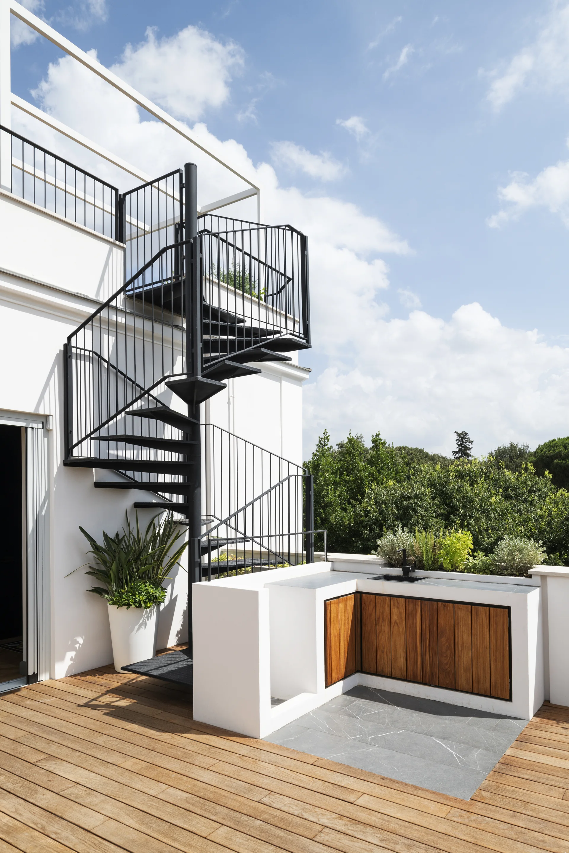A sunny rooftop terrace featuring a black metal spiral staircase, wooden decking, and a built-in kitchen with wood cabinetry.