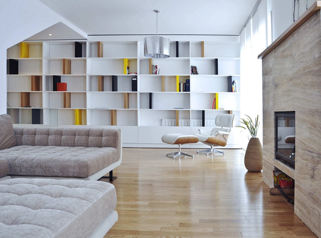 A spacious living room features a tufted beige sofa, a white lounge chair, and a large white built-in shelving unit with colored accent dividers.