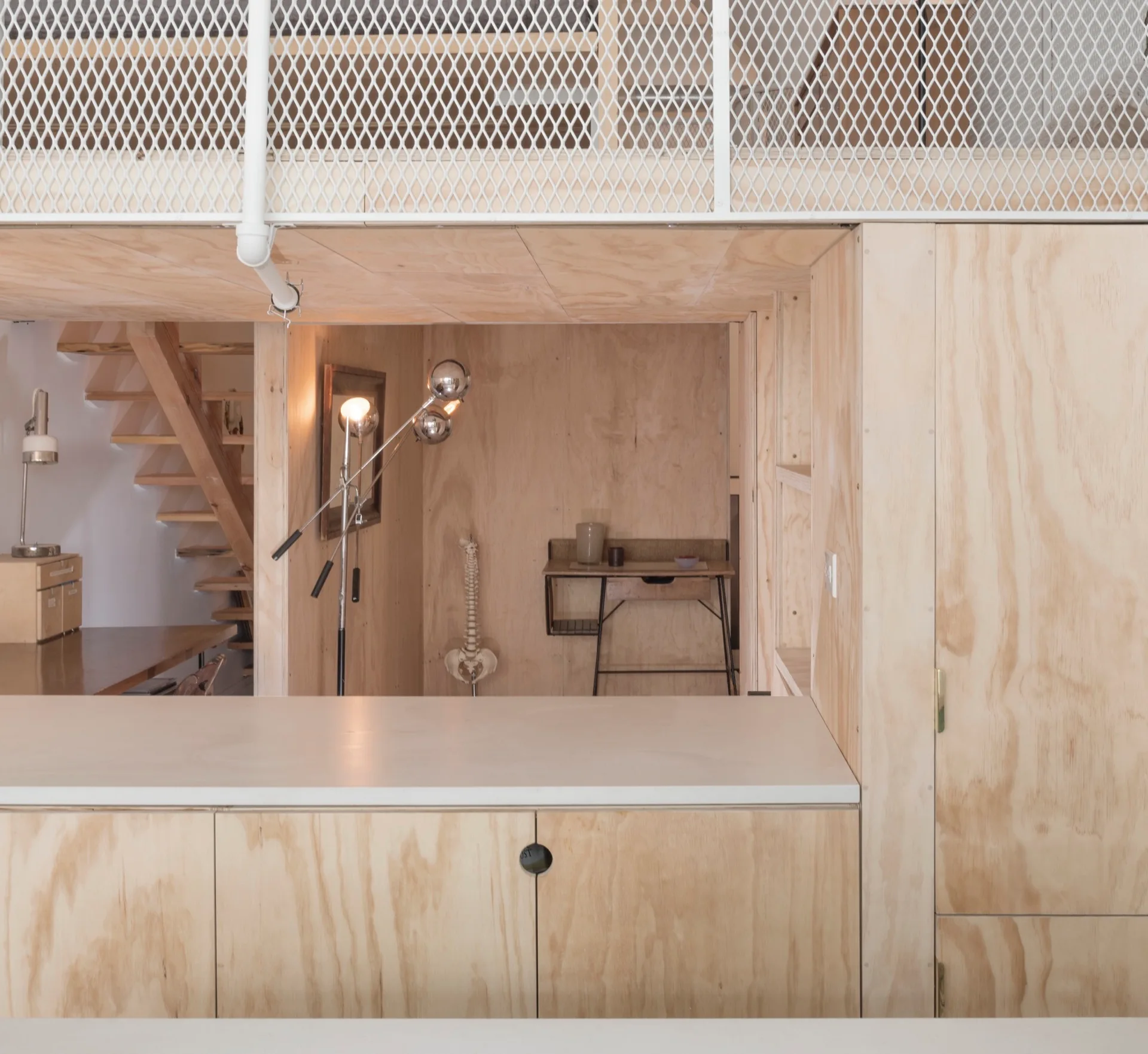 A minimalist split-level loft featuring plywood wall panels, built-in cabinetry, and a white metal mesh railing on the mezzanine.