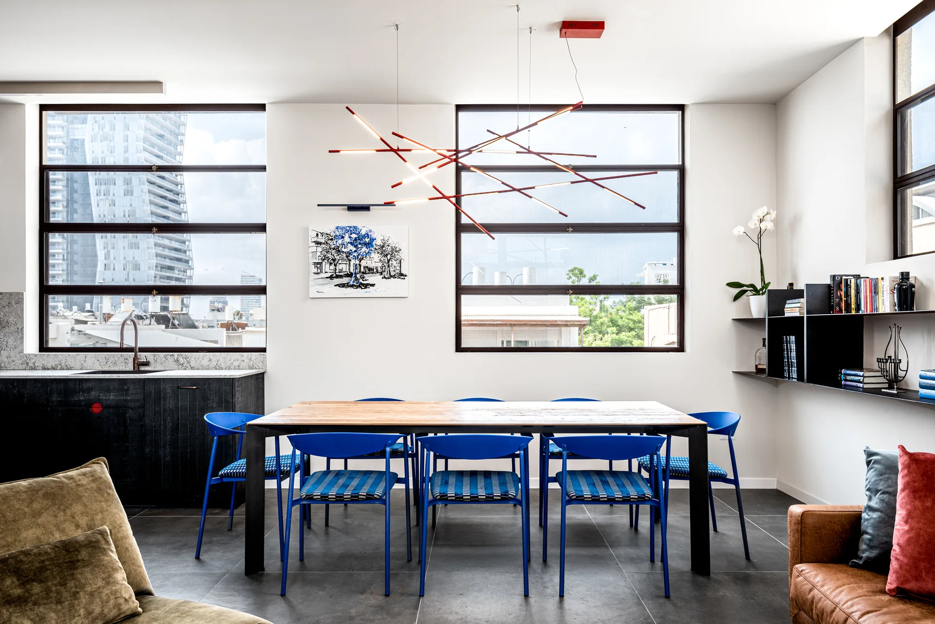A wooden dining table surrounded by bright blue chairs stands under a geometric branch chandelier next to large black-framed windows.