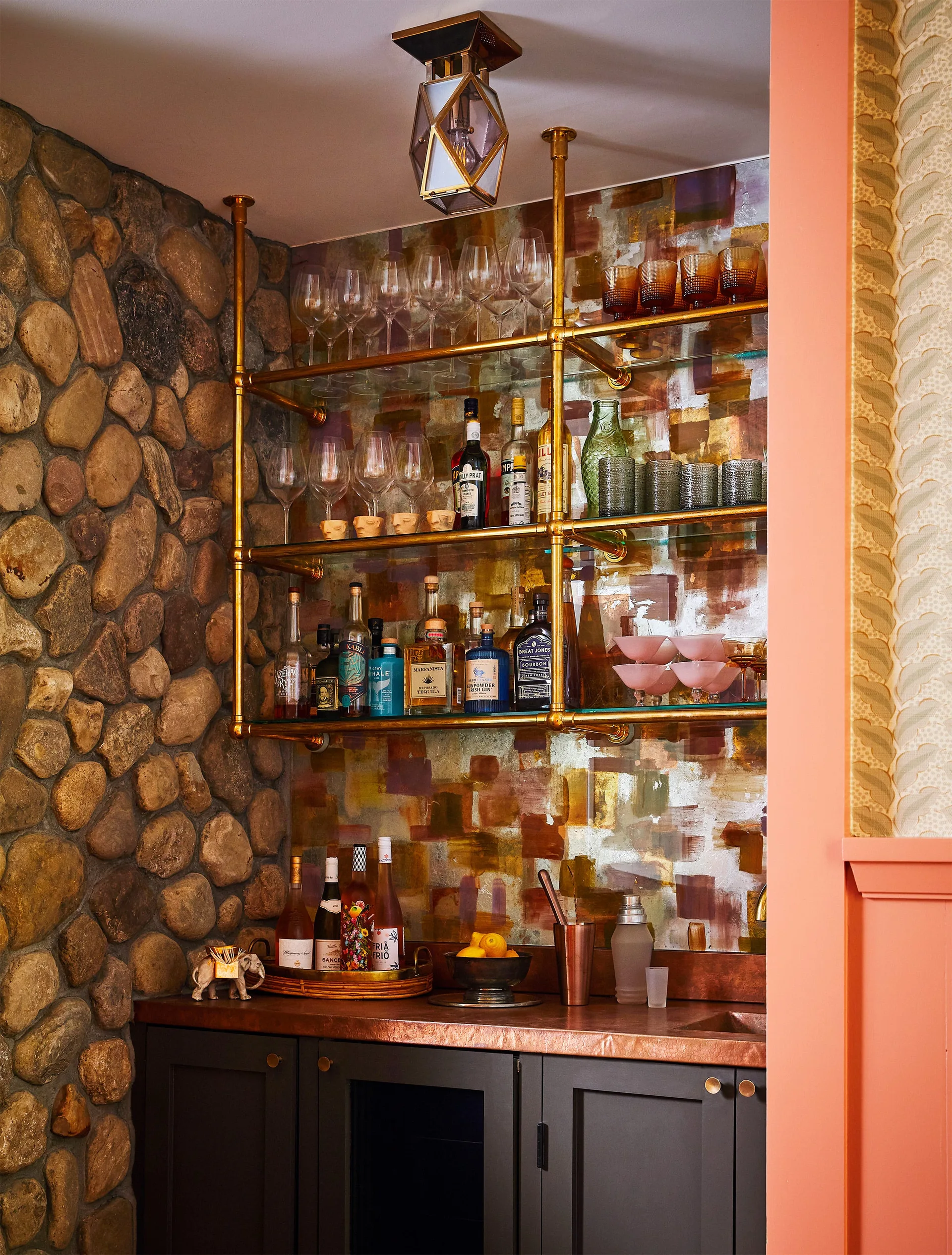 A home bar nook featuring a copper countertop, dark lower cabinets, a brass pipe shelving unit, an abstract backsplash, and a rustic river stone wall.