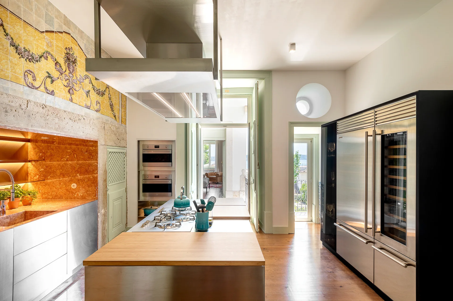 A modern kitchen features a stainless steel island with wood countertops, sage green door frames, and an ornate yellow tile accent wall.