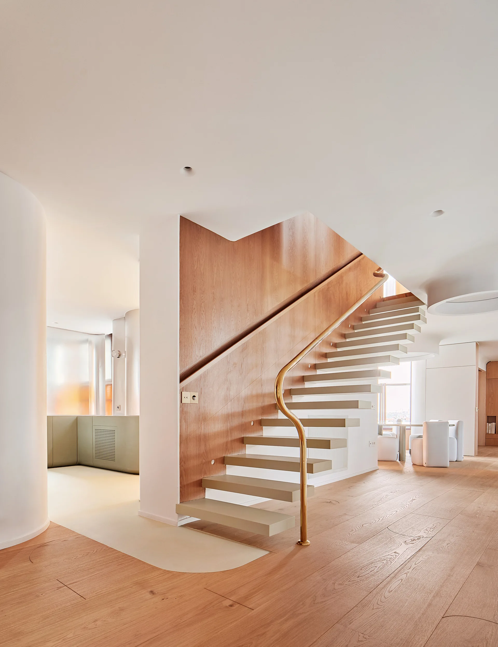 An open-riser staircase features beige treads, a curved brass handrail, and a warm wood-paneled accent wall in a minimalist, light-filled interior.