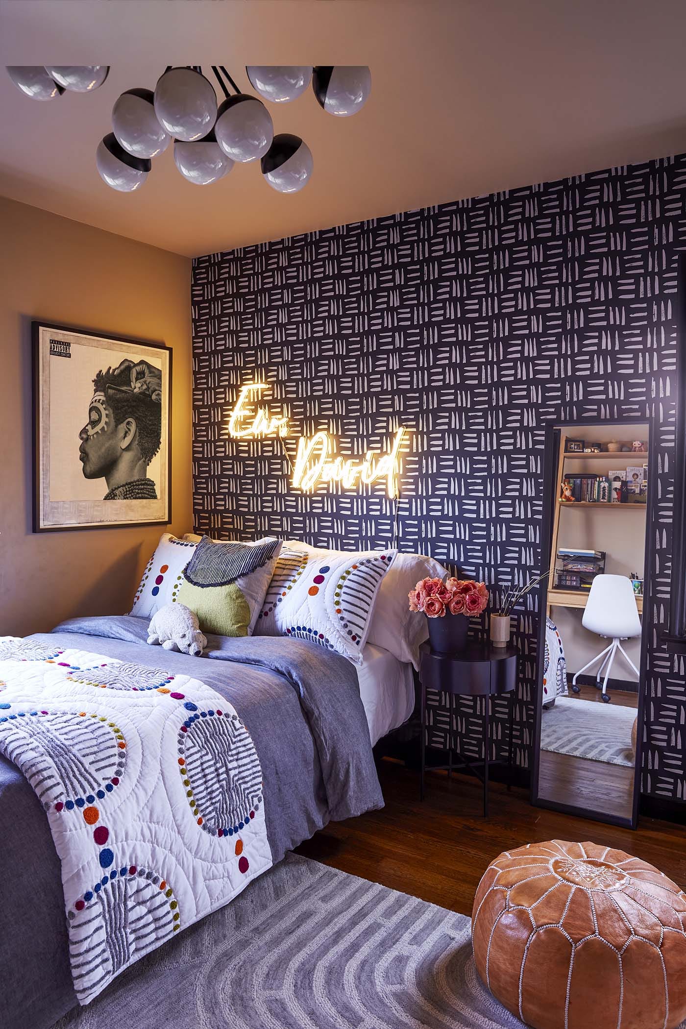 An eclectic bedroom featuring a black graphic patterned accent wall with a glowing neon sign above the bed, a colorful quilt, and a brown leather pouf.