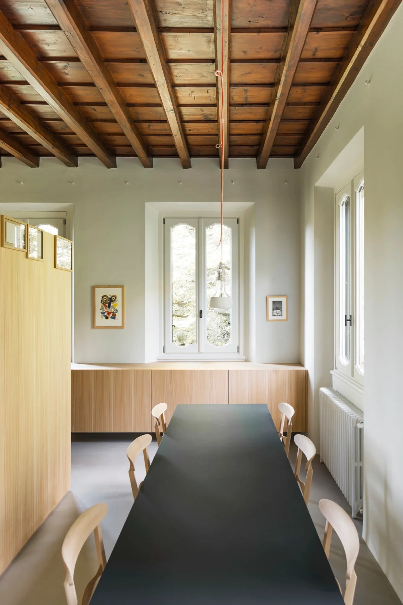 A black dining table and light wood chairs stand beneath an exposed wood beam ceiling next to low wooden cabinets.