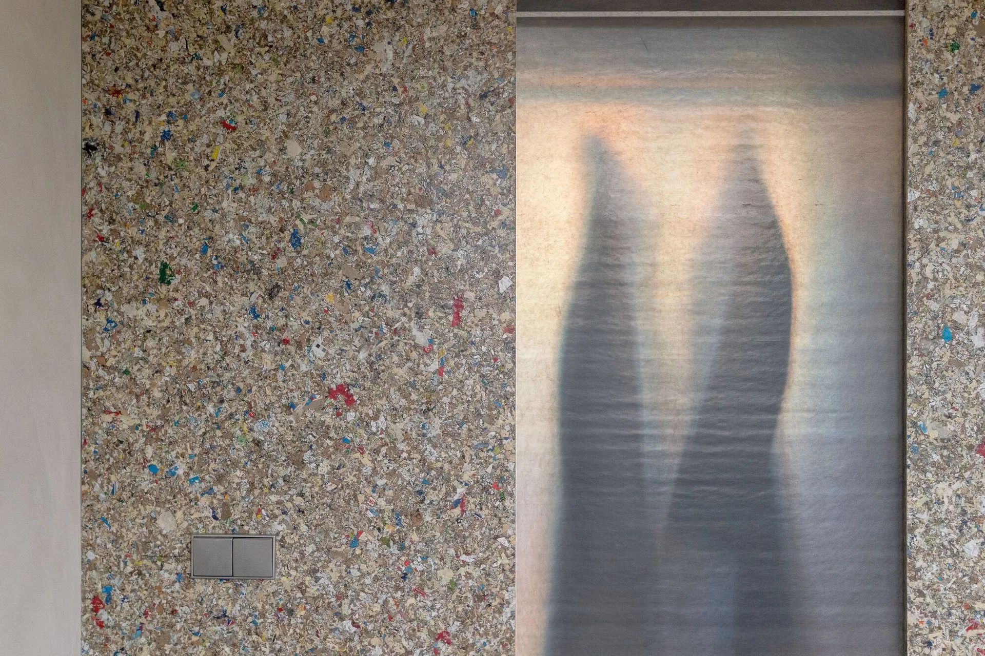 A textured wall made of multi-colored recycled composite material sits next to a glowing, backlit translucent panel with soft shadows.