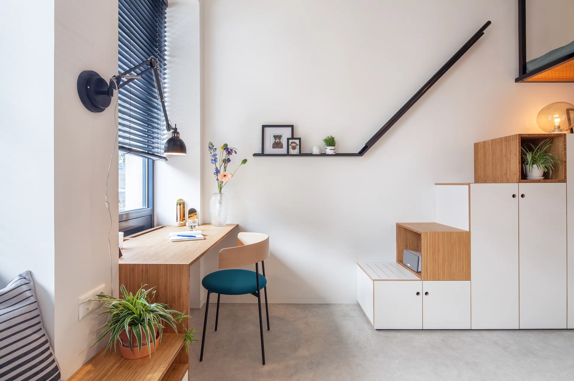 A minimalist home office features a light wood desk, a modular stepped storage cabinet, and an adjustable black wall-mounted task lamp.