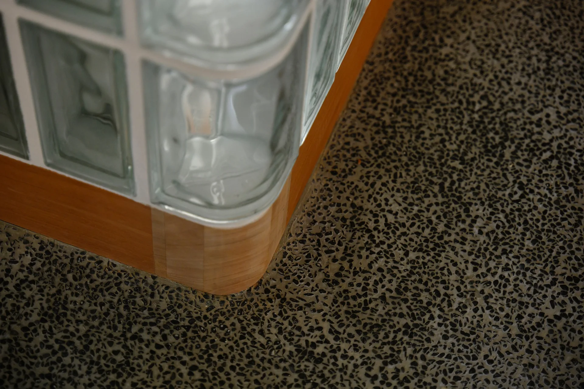 A close-up view of a curved glass block partition with a light wood baseboard on a dark speckled terrazzo floor.