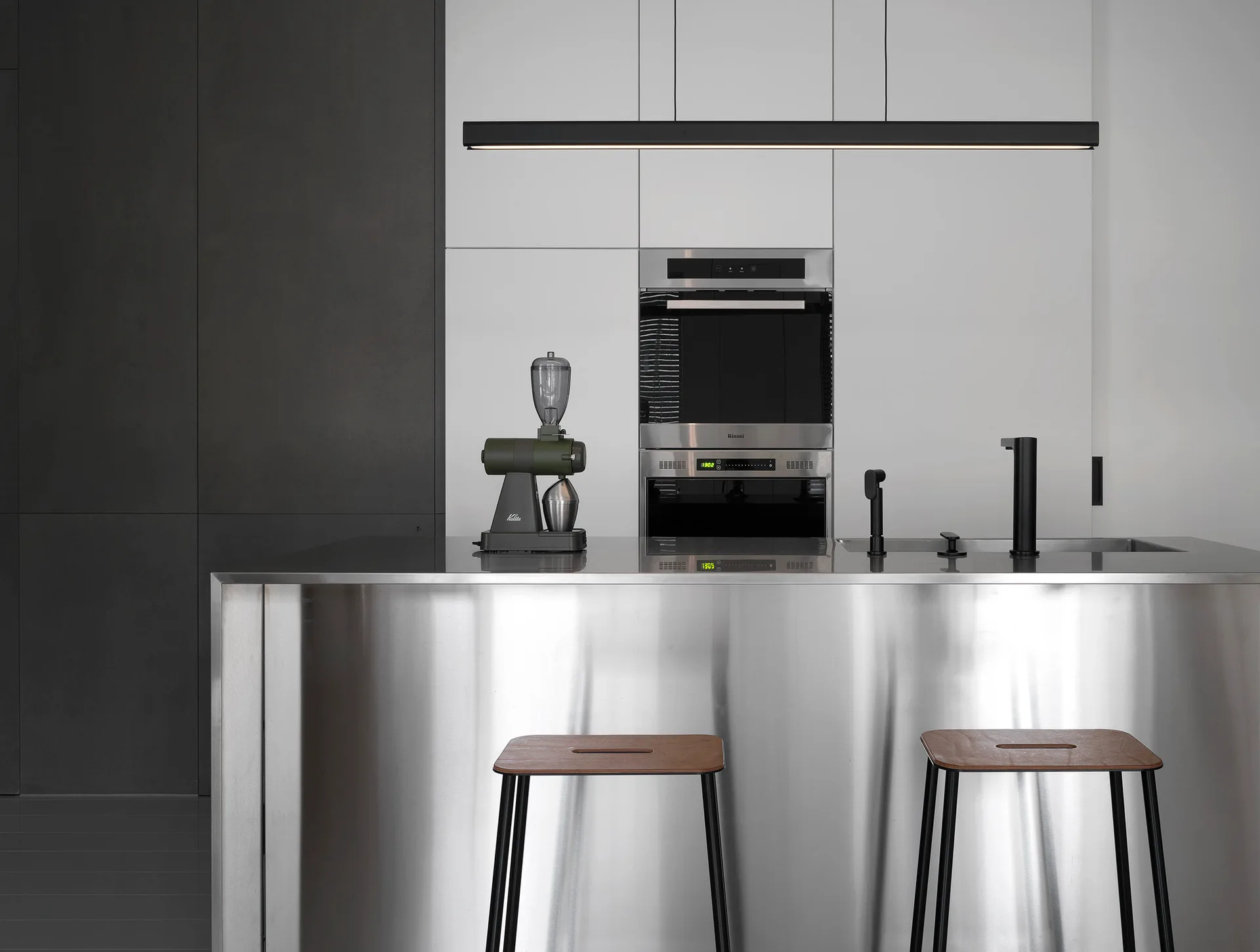 A stainless steel kitchen island with wood-topped bar stools sits in front of flat-panel white cabinets with an integrated oven.