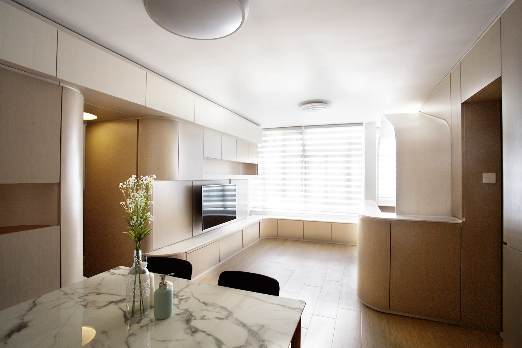 A minimalist living area features custom light-beige wall cabinetry with curved corners, a built-in television, and a veined dining table in the foreground.