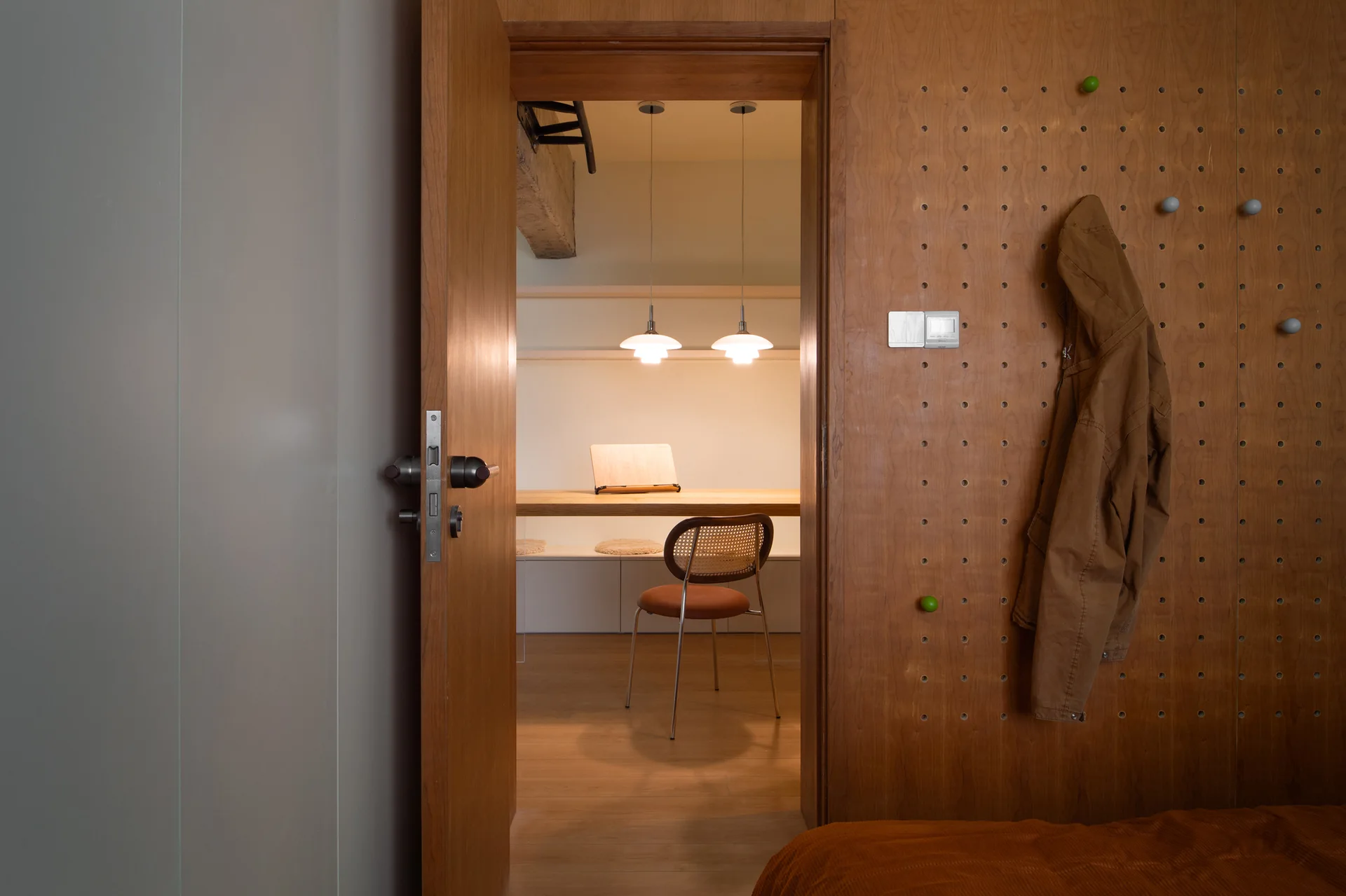 A view through an open wooden doorway reveals a home office featuring a desk, a rattan chair under pendant lights, and an adjacent pegboard wall.