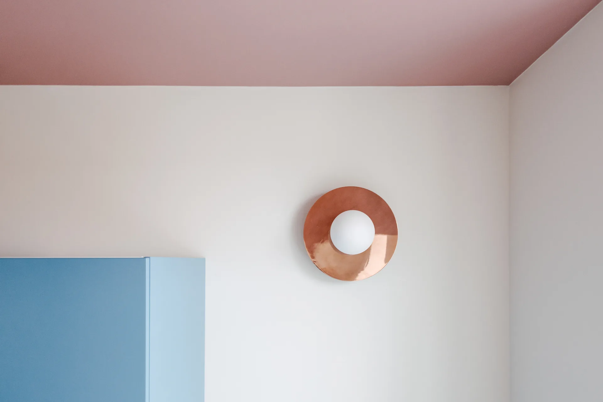 A circular copper wall sconce with a round white bulb is mounted on a white wall below a pink ceiling and next to a blue cabinet.