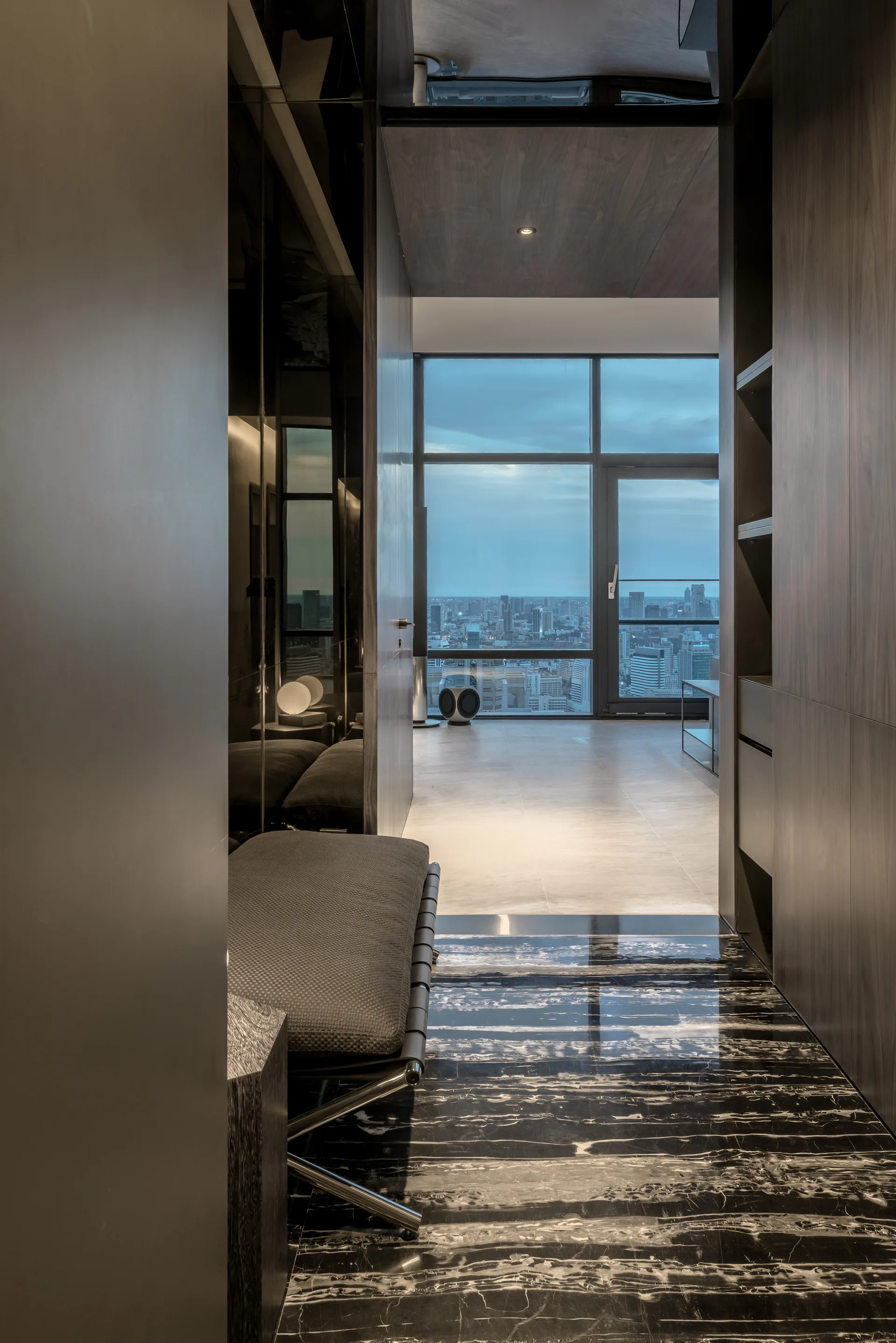 A modern entryway featuring polished black veined marble flooring, wood paneling, a tufted bench, and a distant city view through a large window.