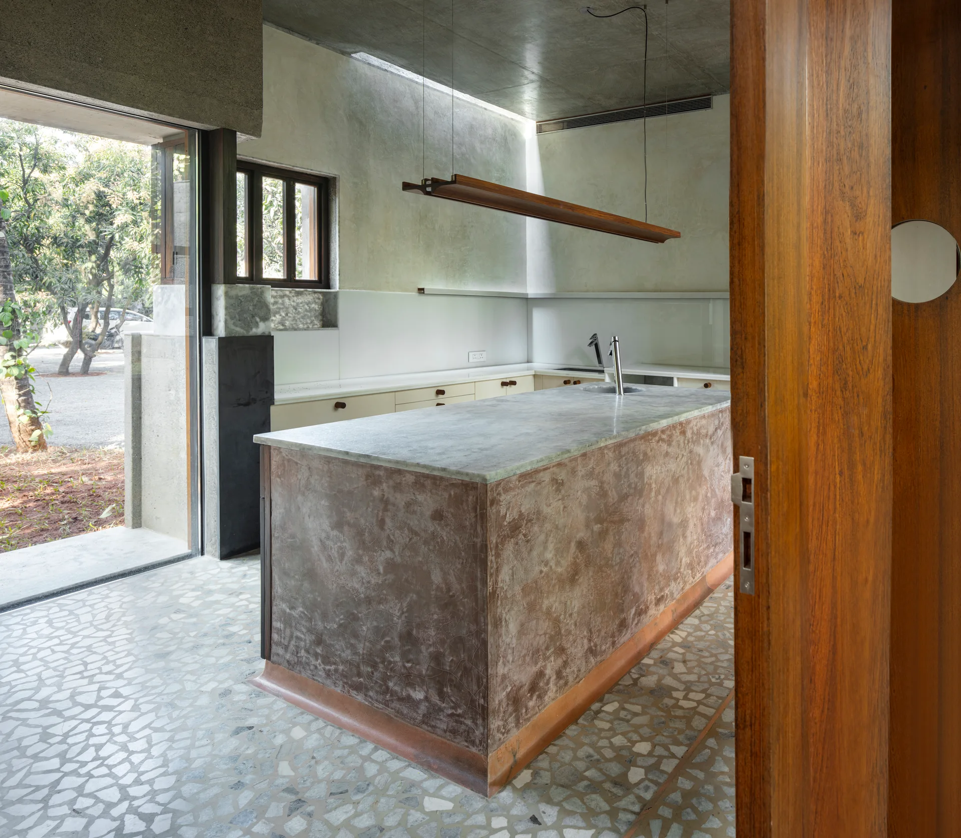 A minimalist kitchen features a textured stone island, light terrazzo flooring, concrete ceiling, and a wooden pendant light next to an outdoor view.