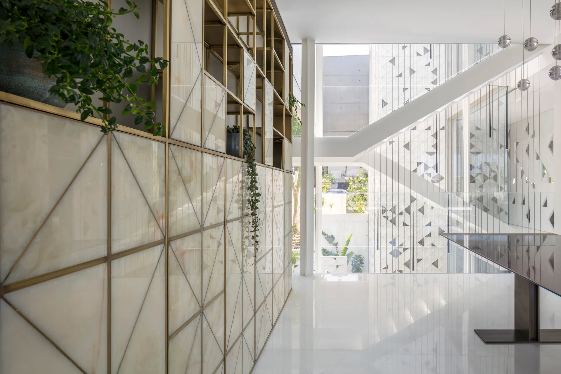 A bright modern interior featuring a large stone-paneled cabinet with a bronze geometric frame, a polished white floor, and a glass-walled staircase.