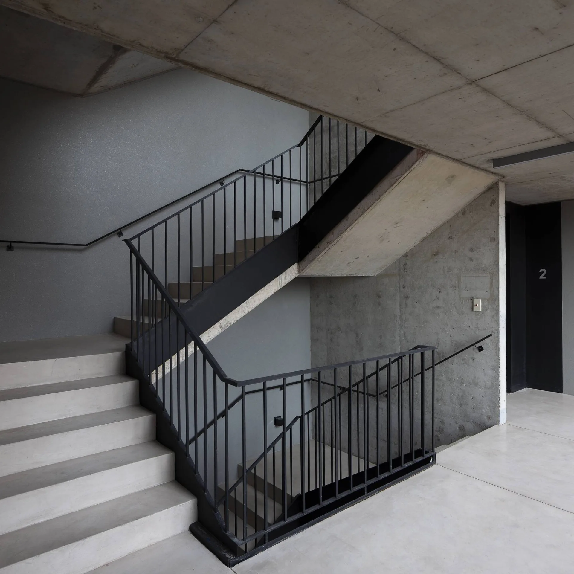 A modern concrete staircase with black metal vertical railings rises through an industrial stairwell with raw concrete walls and ceilings.
