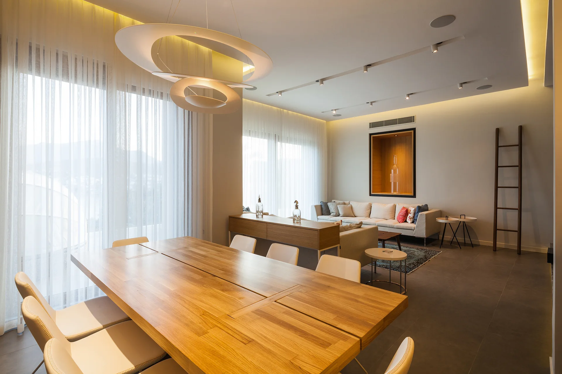 A modern open-plan dining and living space features a large wooden table under a ring pendant light, with a sofa and sheer curtains in the background.
