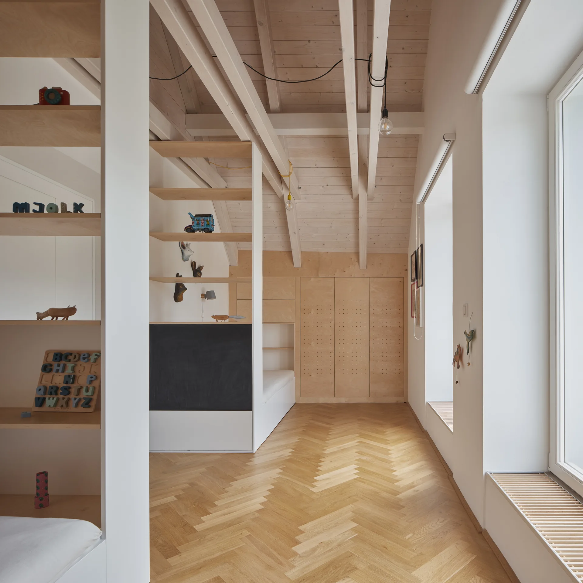 A bright minimalist kids room features light plywood built-in shelving, an integrated chalkboard, and light wood herringbone parquet flooring.