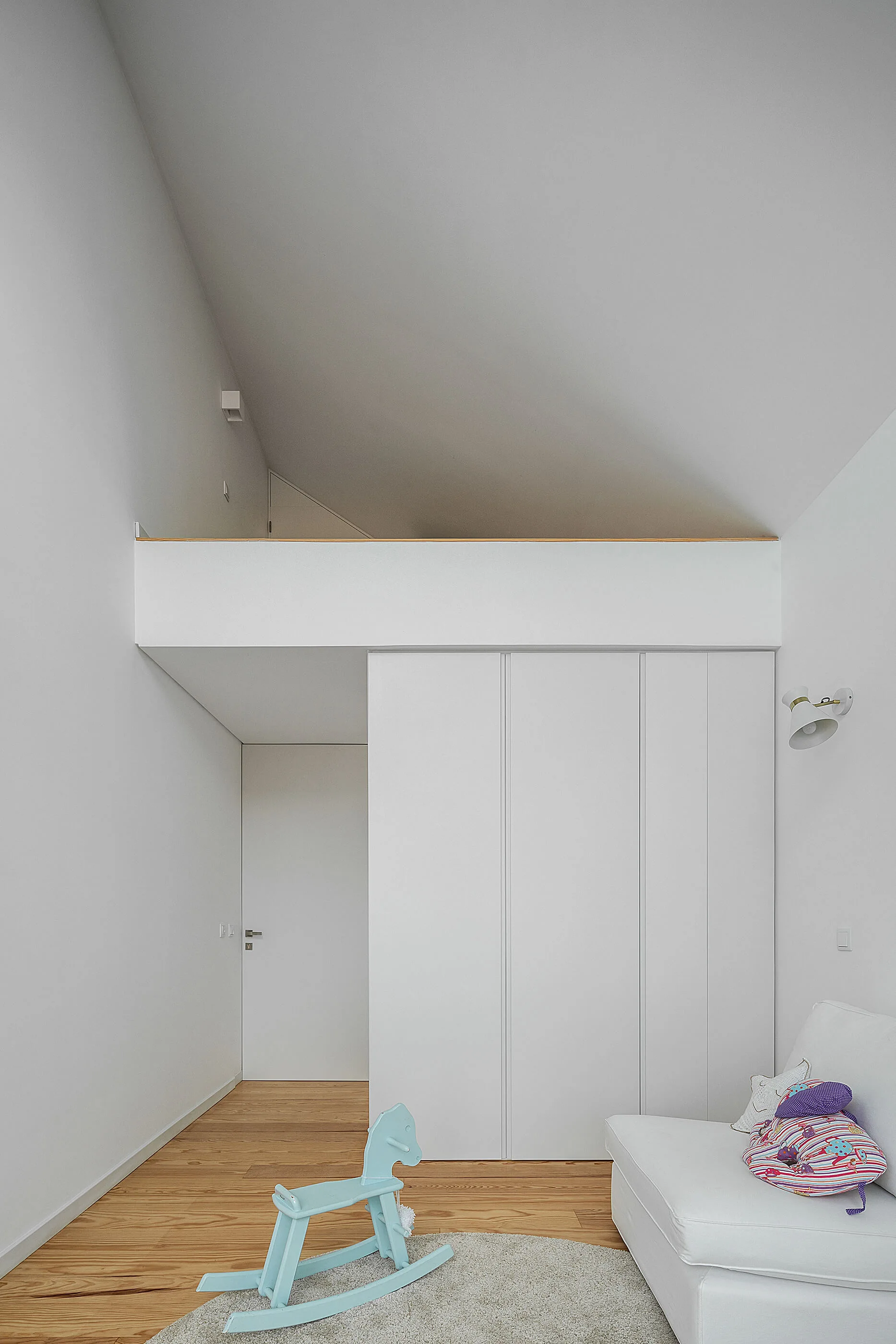 A minimalist white playroom features built-in wardrobes under a lofted platform, light wood floors, and a light blue rocking horse.
