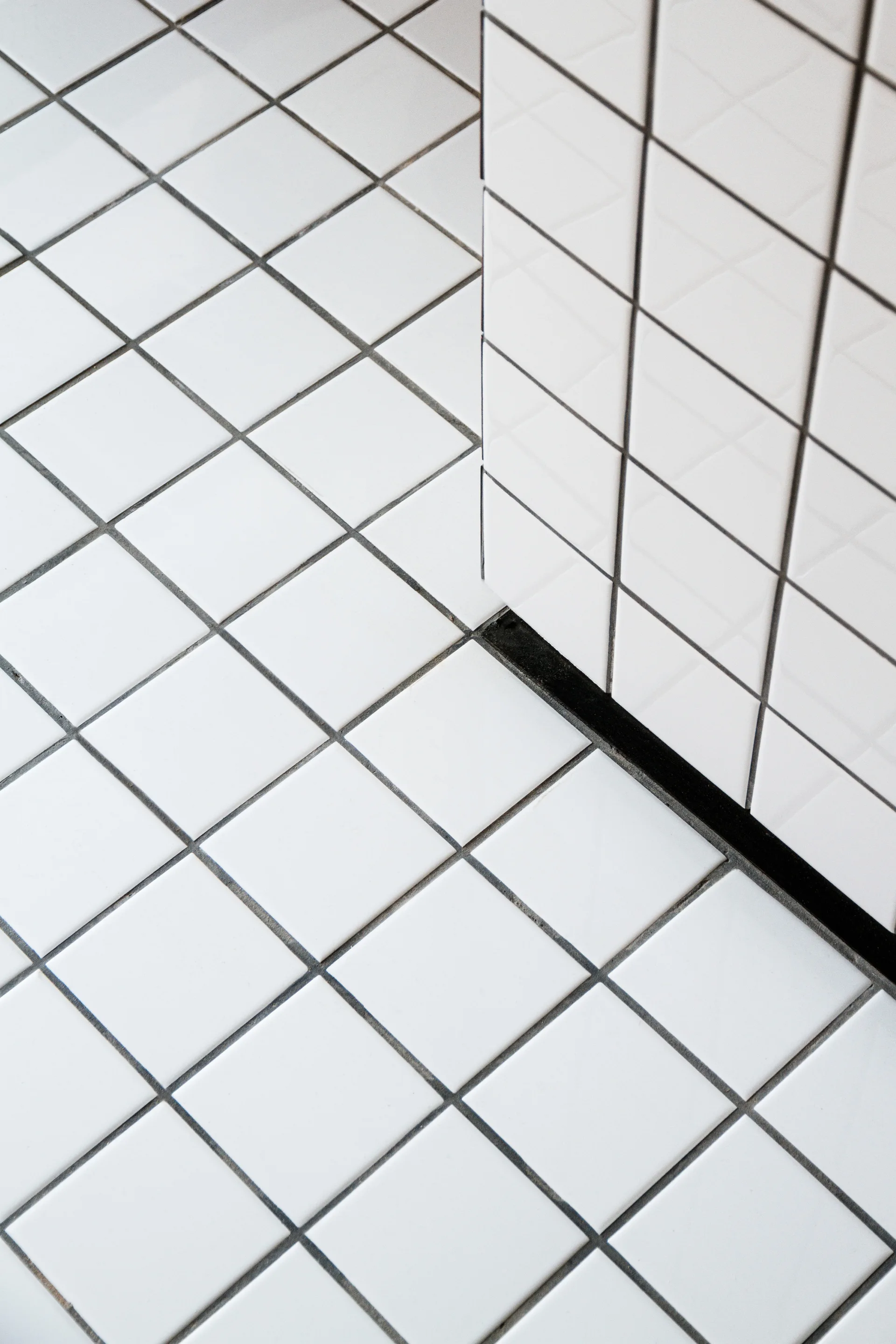 Close-up view of a floor and wall corner clad in glossy white square ceramic tiles with high-contrast dark grout lines.