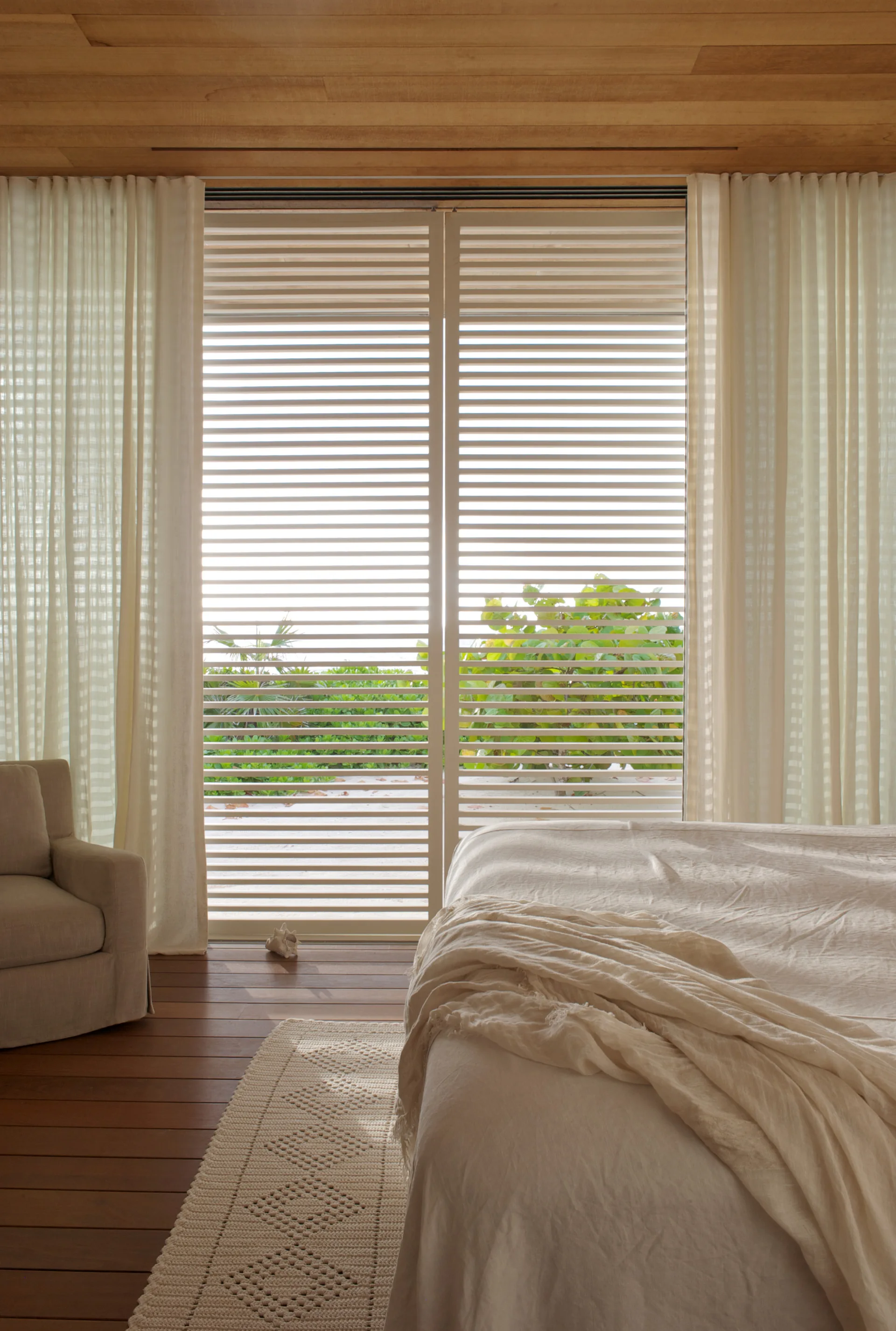A bedroom with a wood plank ceiling and floor features a bed with cream bedding, a beige armchair, and large louvered sliding doors.