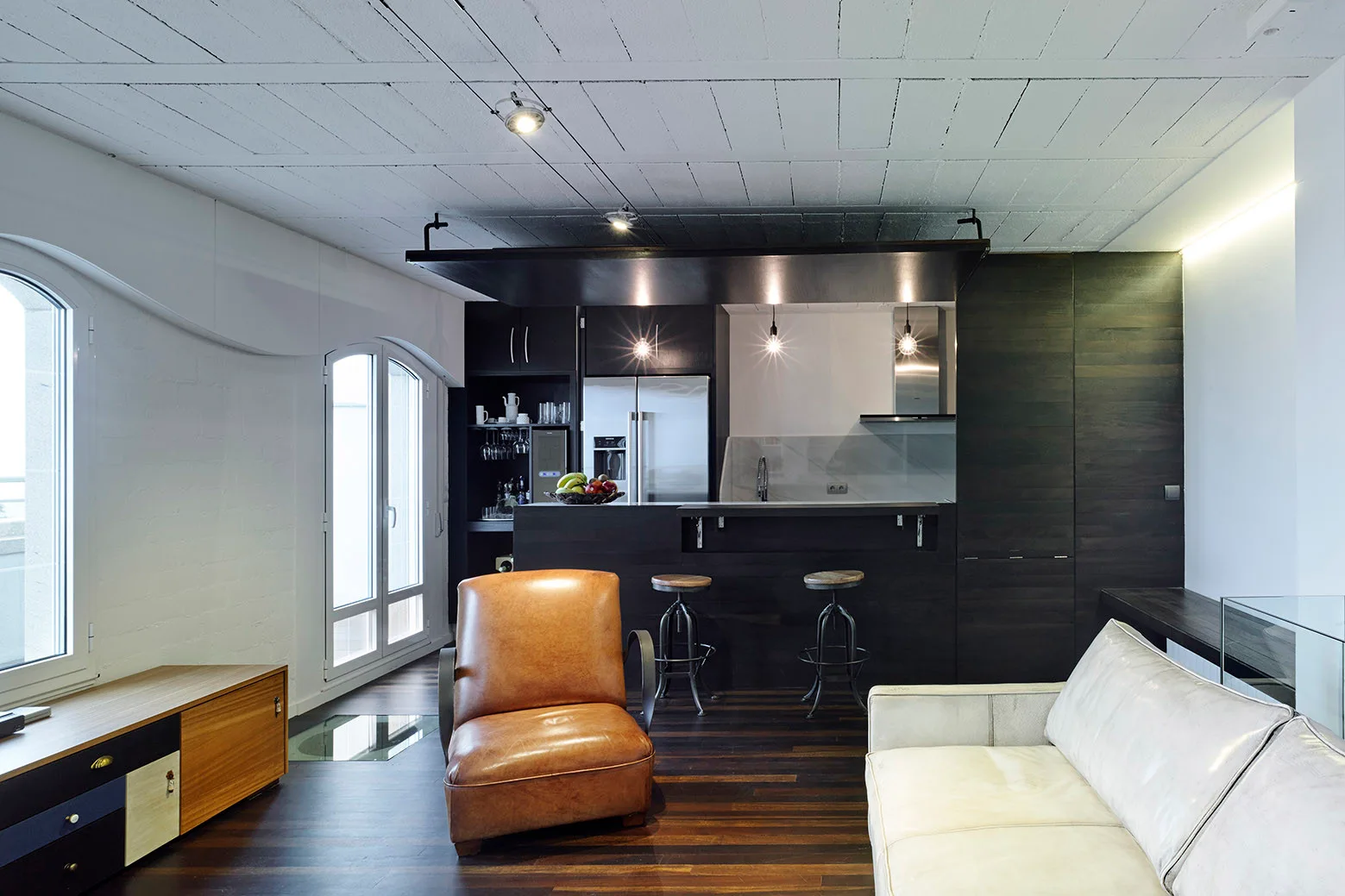 An open-plan living room and kitchen featuring a dark wood island, a tan leather armchair, a light-colored sofa, and a white paneled ceiling.
