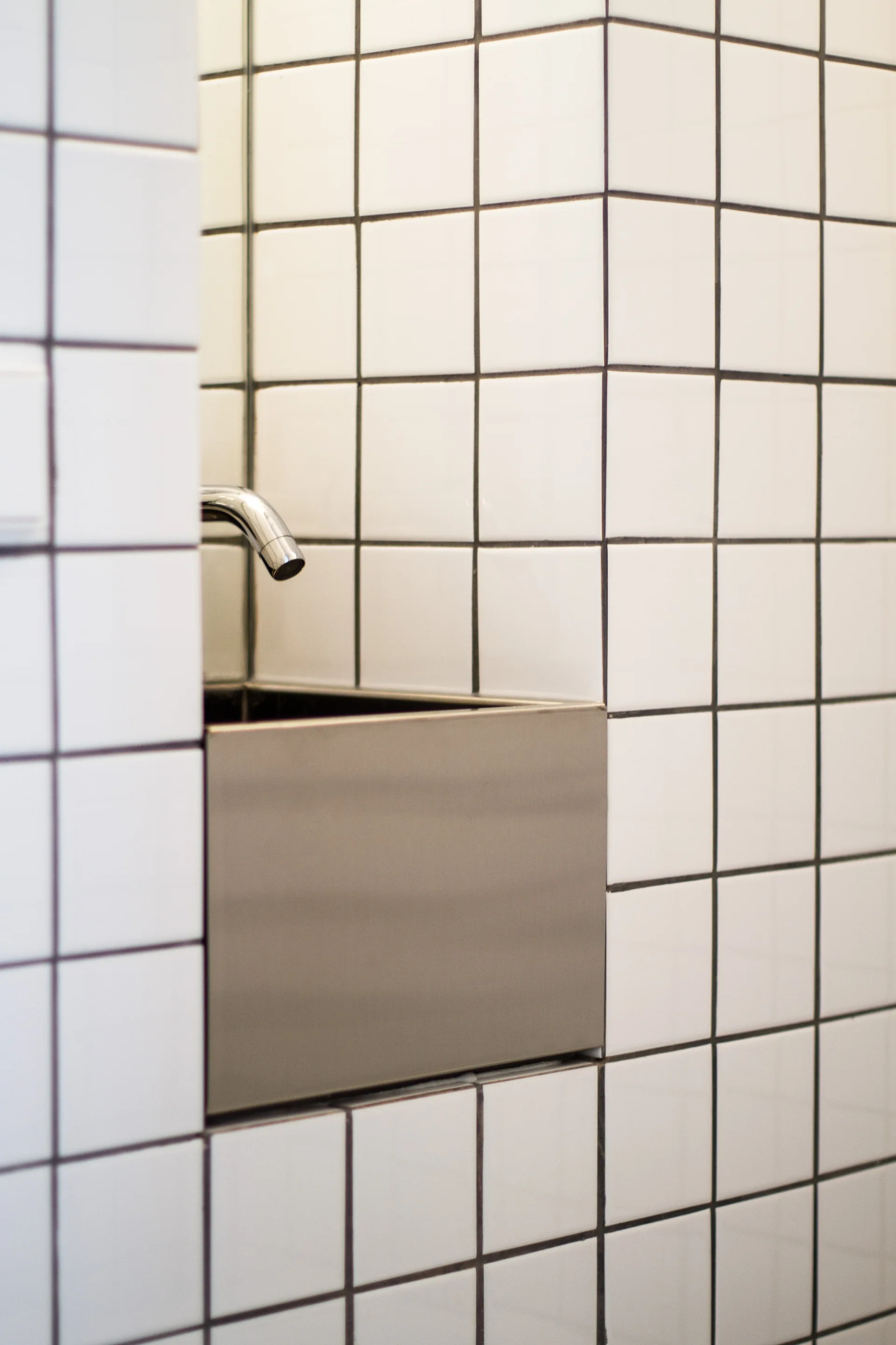 A minimalist metal basin sink and chrome faucet are recessed into a white tiled wall with dark square grout.