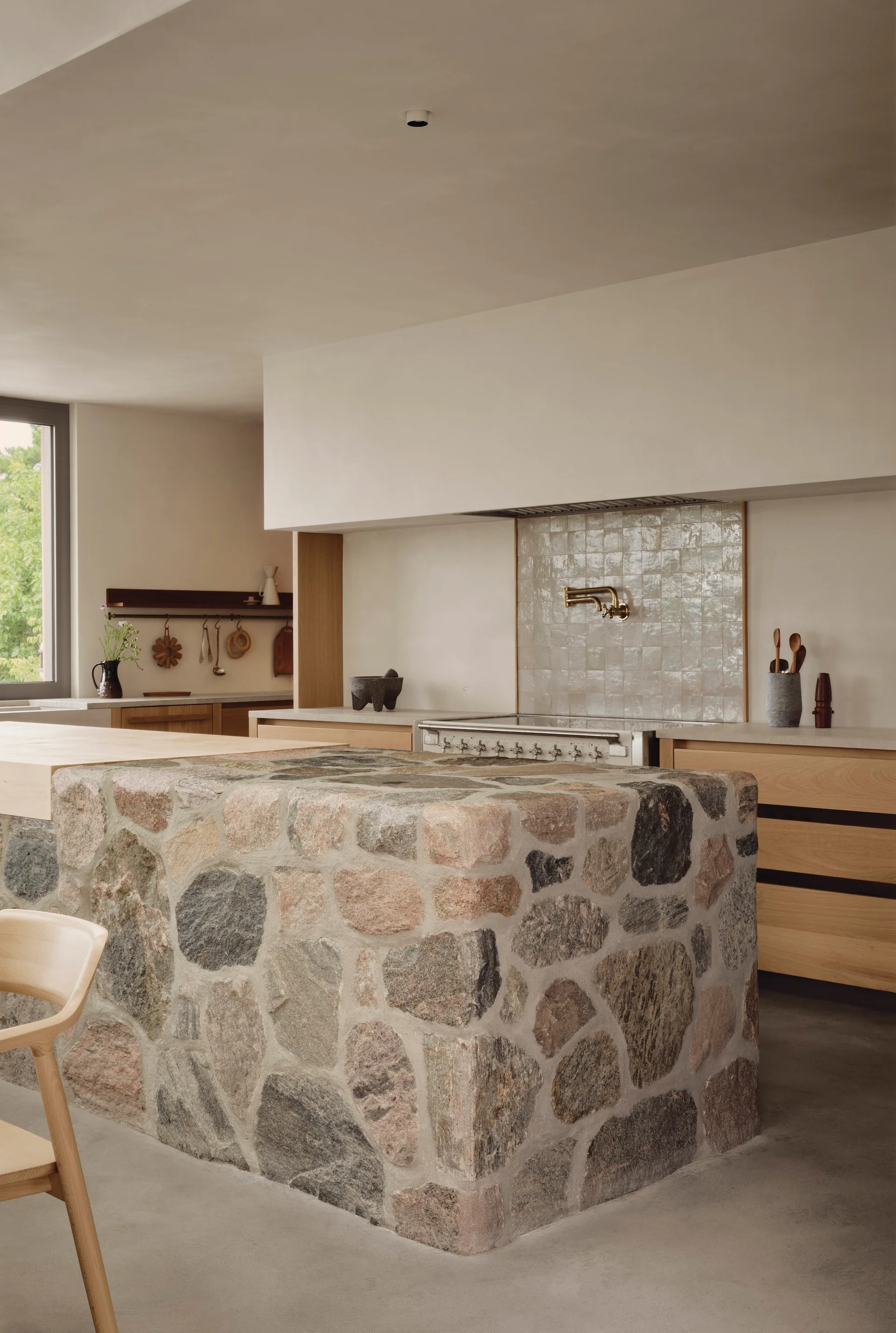 A minimalist kitchen featuring a large stone-clad island, light oak wood cabinetry, and a glazed square tile backsplash behind a stainless steel stove.