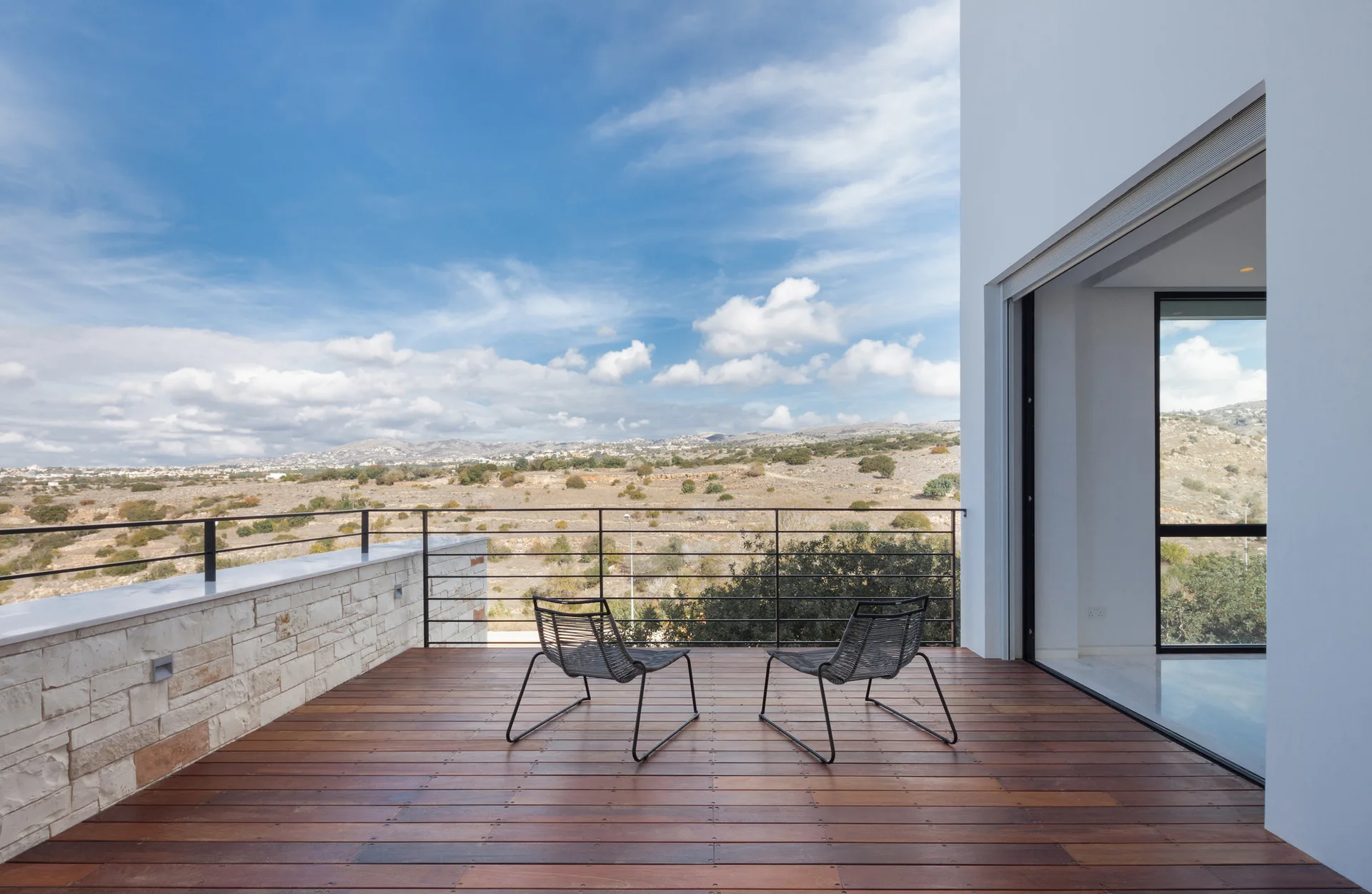 Two minimalist black metal chairs sit on a dark wood plank terrace with a metal railing overlooking a grassy valley landscape.