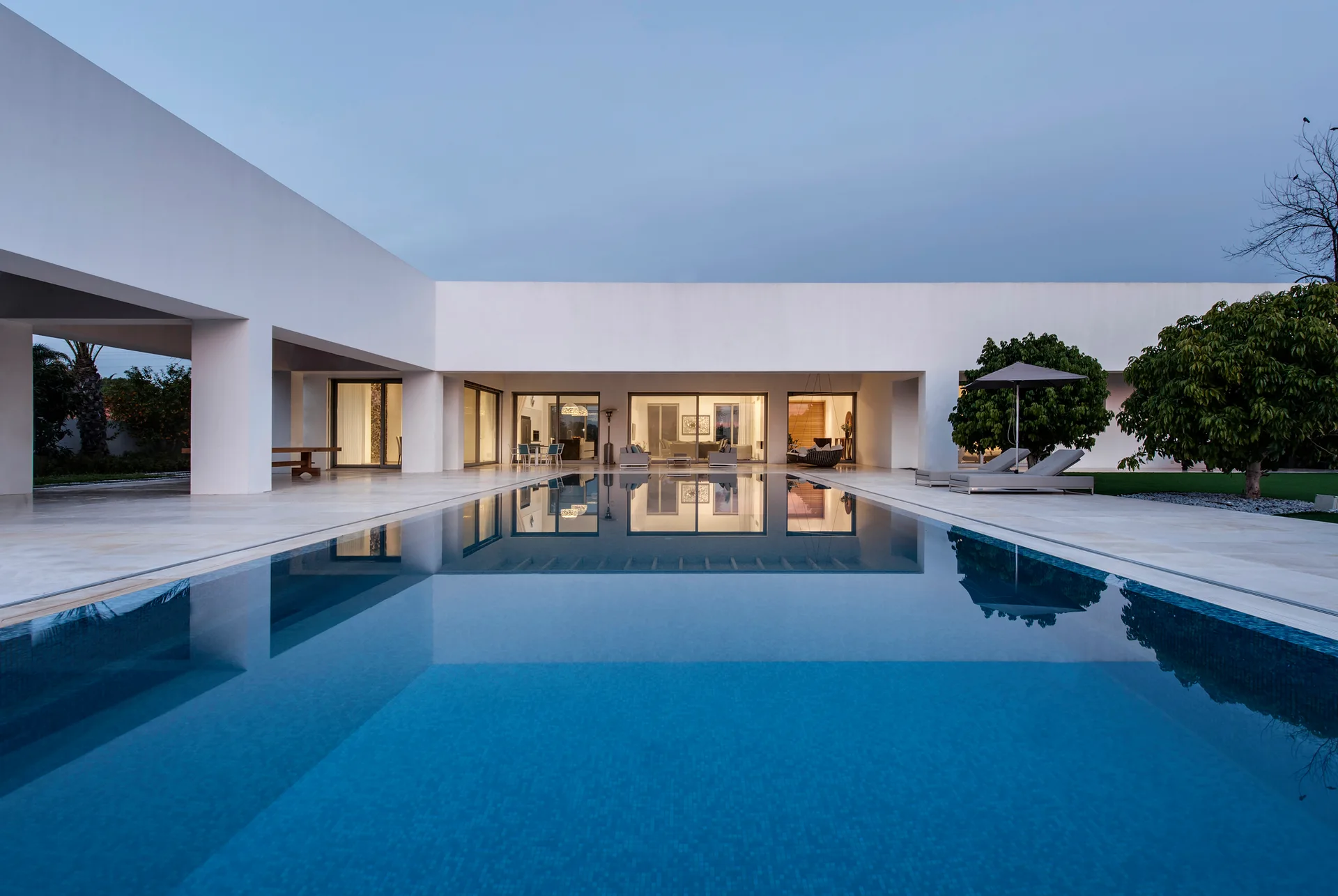 A large rectangular swimming pool reflects a modern white stucco house with glass sliding doors and a stone tiled patio at dusk.