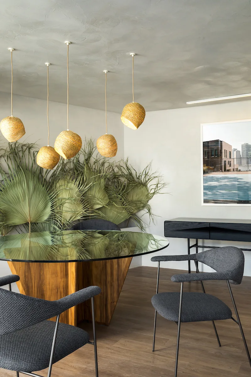 A modern dining room features a round wood and glass table, grey upholstered chairs, woven pendant lights, and large green palm fronds against a grey wall.